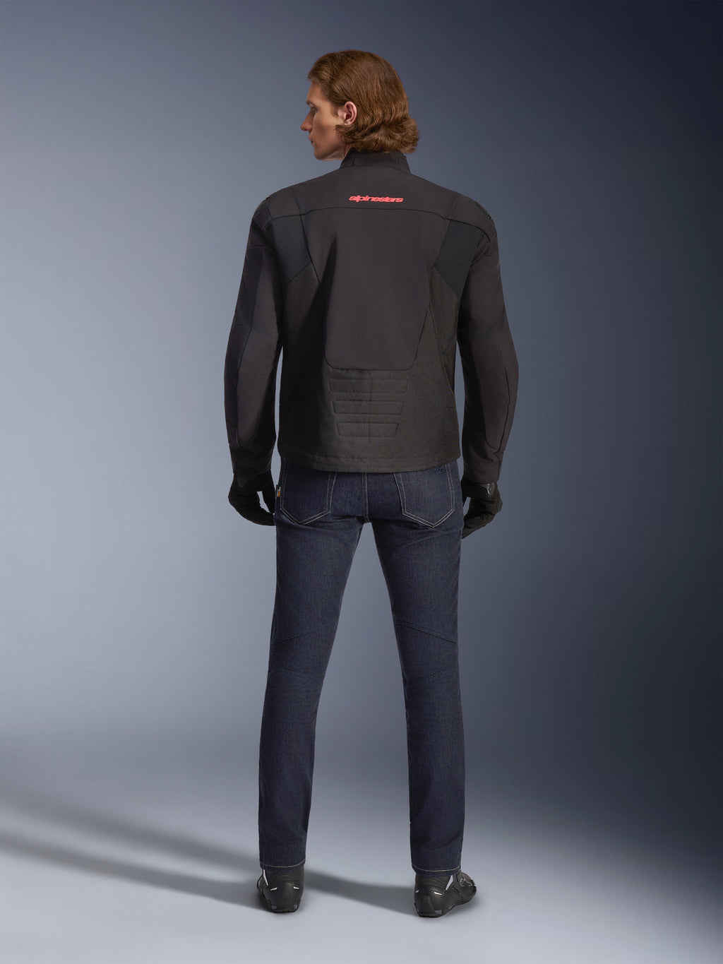 Giacca Alpinestars SMX Waterproof, nero, vista posteriore indossata da un modello, con logo rosso sulla parte superiore della schiena, orlo posteriore allungato per una maggiore copertura durante la guida, abbinata a jeans da moto blu scuro e guanti neri.