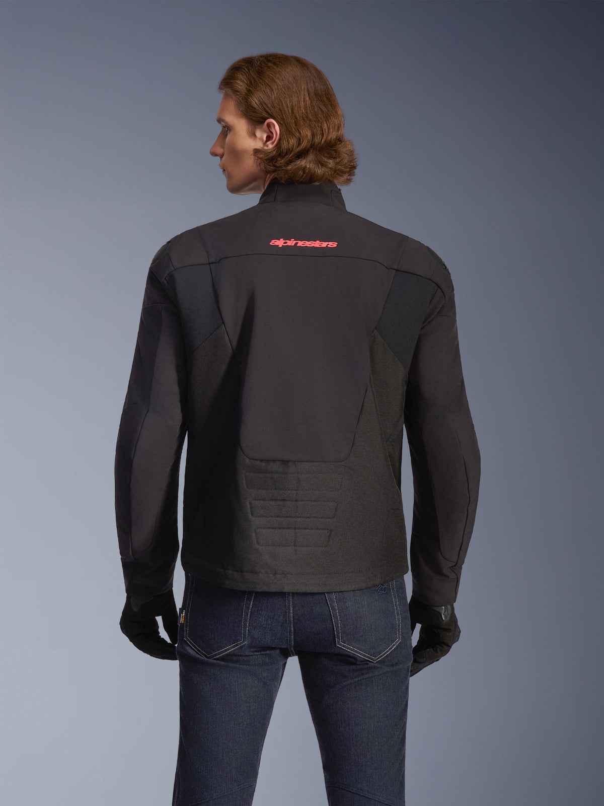 Giacca Alpinestars SMX Waterproof, nera con logo rosso acceso sul retro, vista posteriore su un modello, indossata con jeans blu scuro e guanti neri, giacca da moto in tessuto per guida su strada e sportiva.
