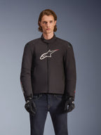 Alpinestars SMX Waterproof Jacket, giacca da moto in tessuto nero con logo a stella bianco e rosso sul petto, indossata da un modello con guanti neri e jeans blu, dotata di cursori esterni sulle spalle e vestibilità sportiva