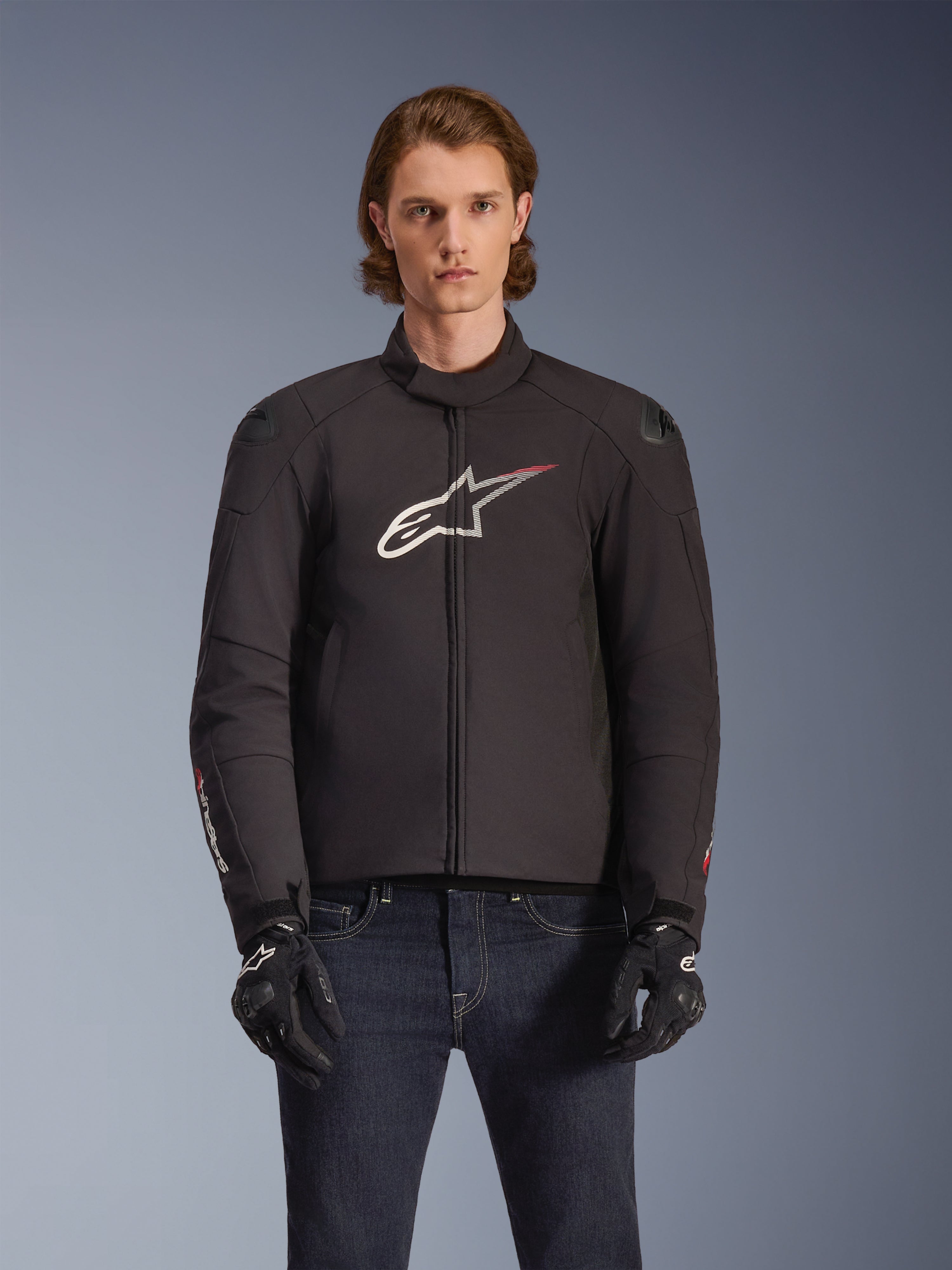 Alpinestars SMX Waterproof Jacket, giacca da moto in tessuto nero con logo a stella bianco e rosso sul petto, indossata da un modello con guanti neri e jeans blu, dotata di cursori esterni sulle spalle e vestibilità sportiva