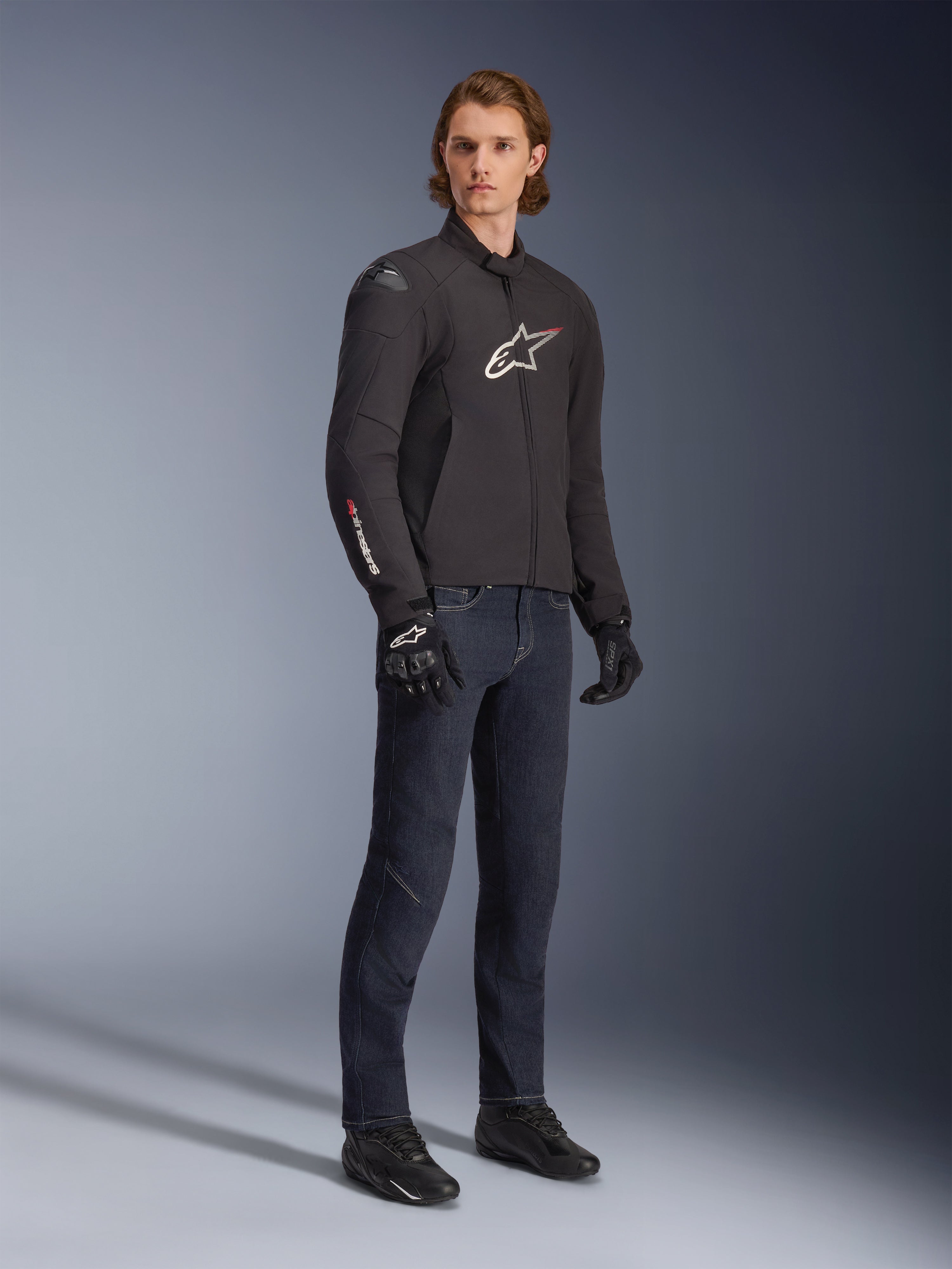 Modello che indossa la giacca Alpinestars SMX Waterproof, nera con accenti del logo bianchi e rossi, abbinata a pantaloni in denim blu scuro e guanti da moto neri, elegante giacca da moto in tessuto con vestibilità sportiva.