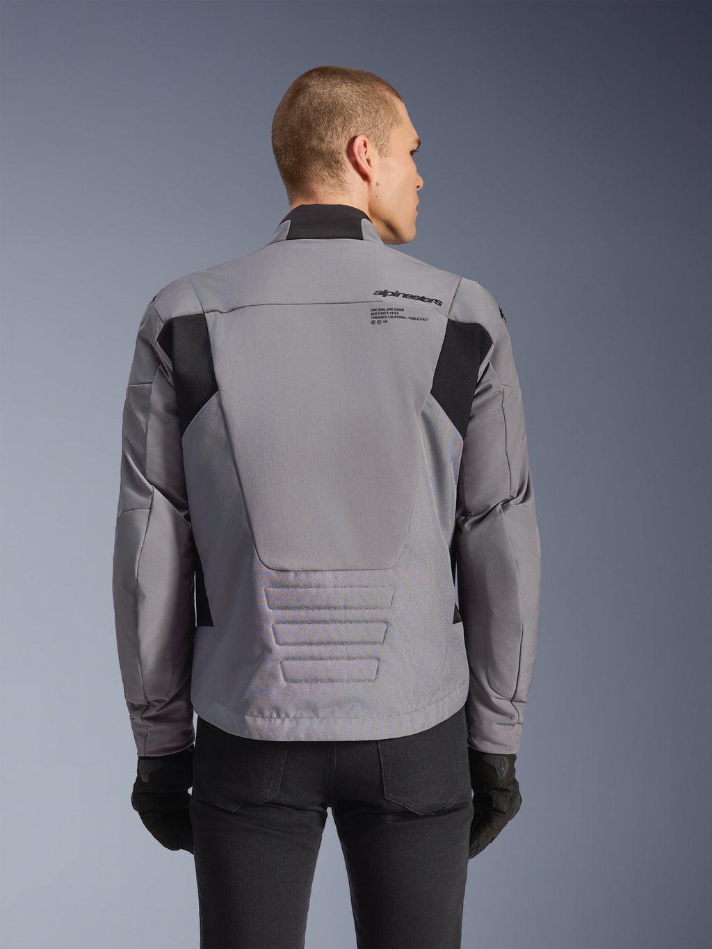 Giacca impermeabile Alpinestars SMX, grigio con accenti neri, vista posteriore che mostra il logo sulla schiena e il profilo posteriore allungato, indossata da un modello con pantaloni e guanti neri