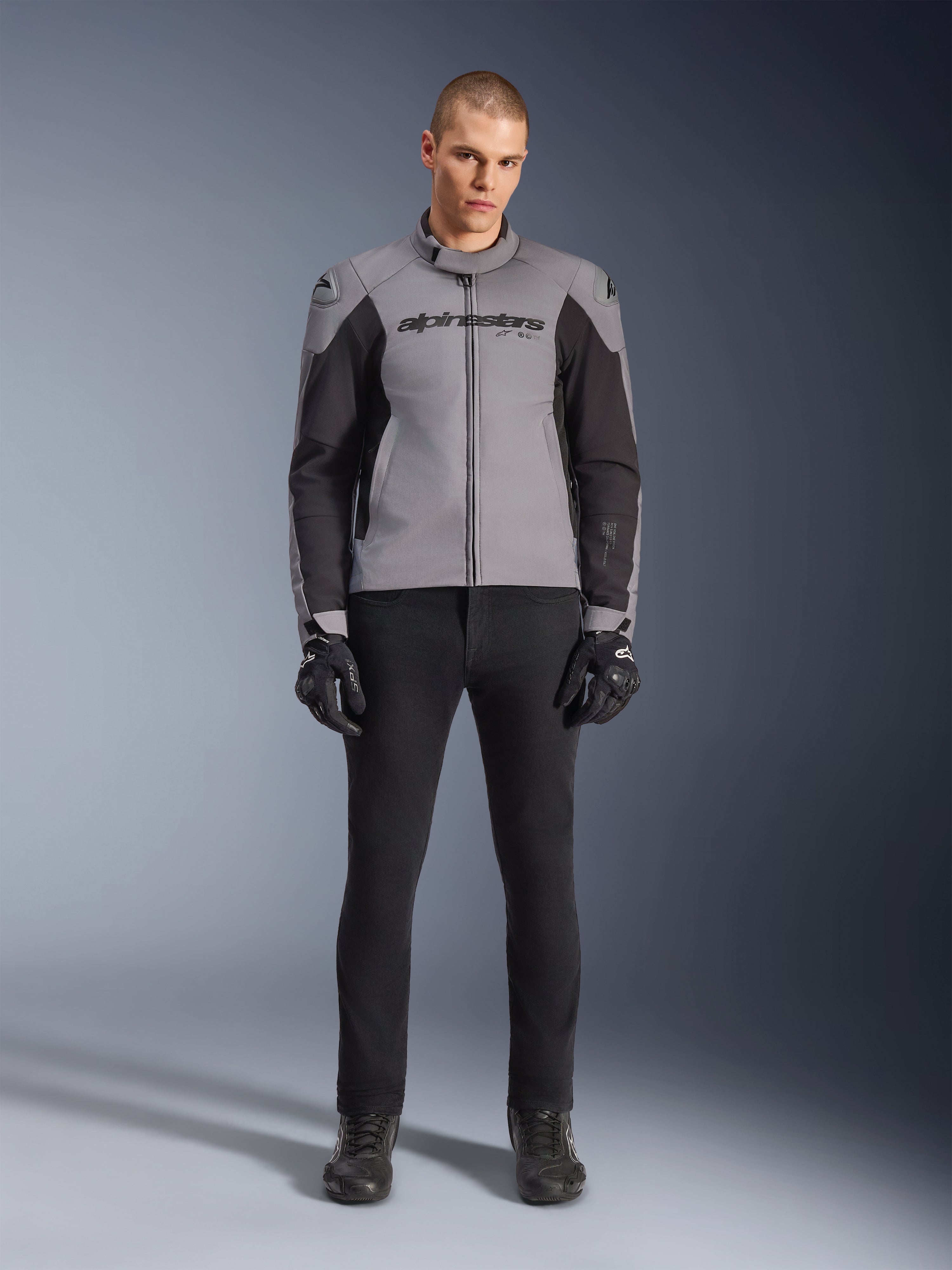 Giacca Alpinestars SMX Waterproof, grigio scuro e nero, vista frontale indossata da un modello, giacca da moto sportiva in tessuto con pannelli laterali neri e marchio Alpinestars sul petto, abbinata a pantaloni da guida e guanti neri.