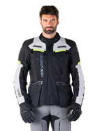 Uomo che indossa la giacca Alpinestars Bogotá Pro Drystar®, nero, grigio ghiaccio e giallo fluo, giacca da moto adventure touring quattro stagioni, design impermeabile con tasche multiple e branding verticale, abbinata a guanti neri e pantaloni da moto grigi