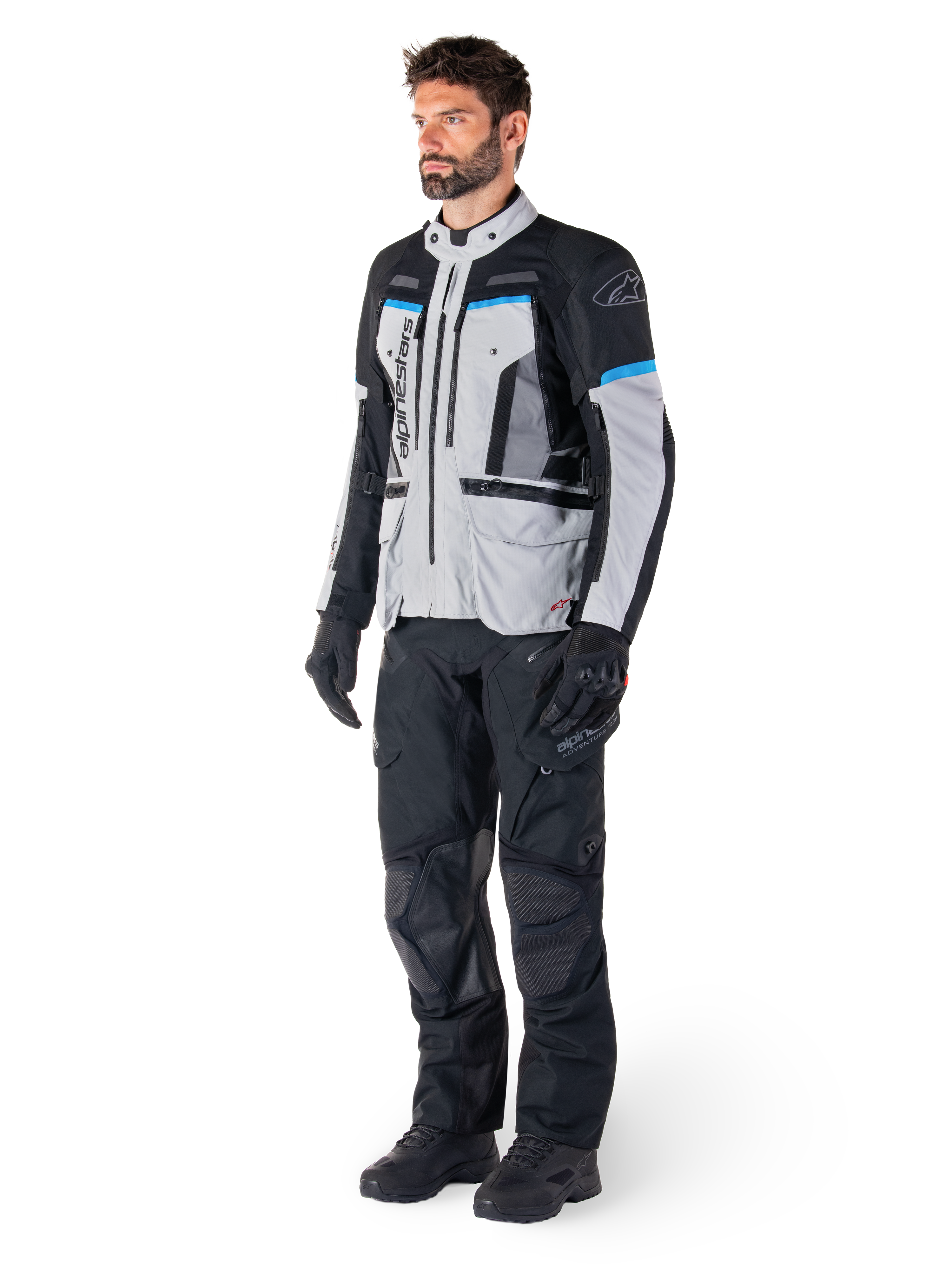 Pilota uomo che indossa la giacca Alpinestars Bogotá Pro Drystar® in Ice Gray, Black e Cyan, abbinata a pantaloni tecnici da guida e guanti neri, equipaggiamento adventure touring per prestazioni in ogni condizione atmosferica.