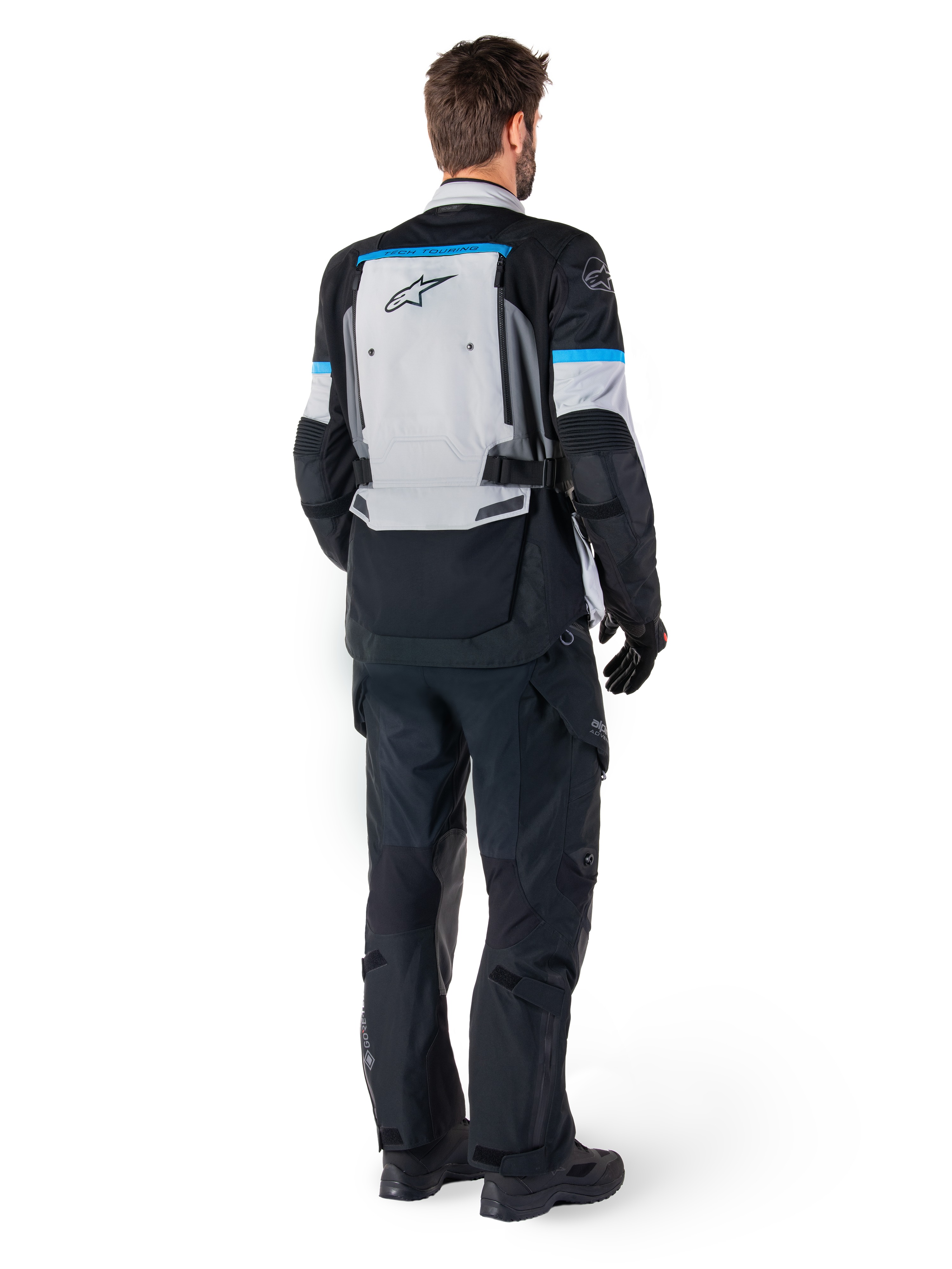 Alpinestars Bogota' Pro Drystar® Jacket, giacca da moto adventure, grigio ghiaccio, nero e ciano, vista posteriore indossata con pantaloni e guanti neri coordinati, caratterizzata da un ampio pannello posteriore grigio con logo Alpinestars nero e dettagli in blu ciano.