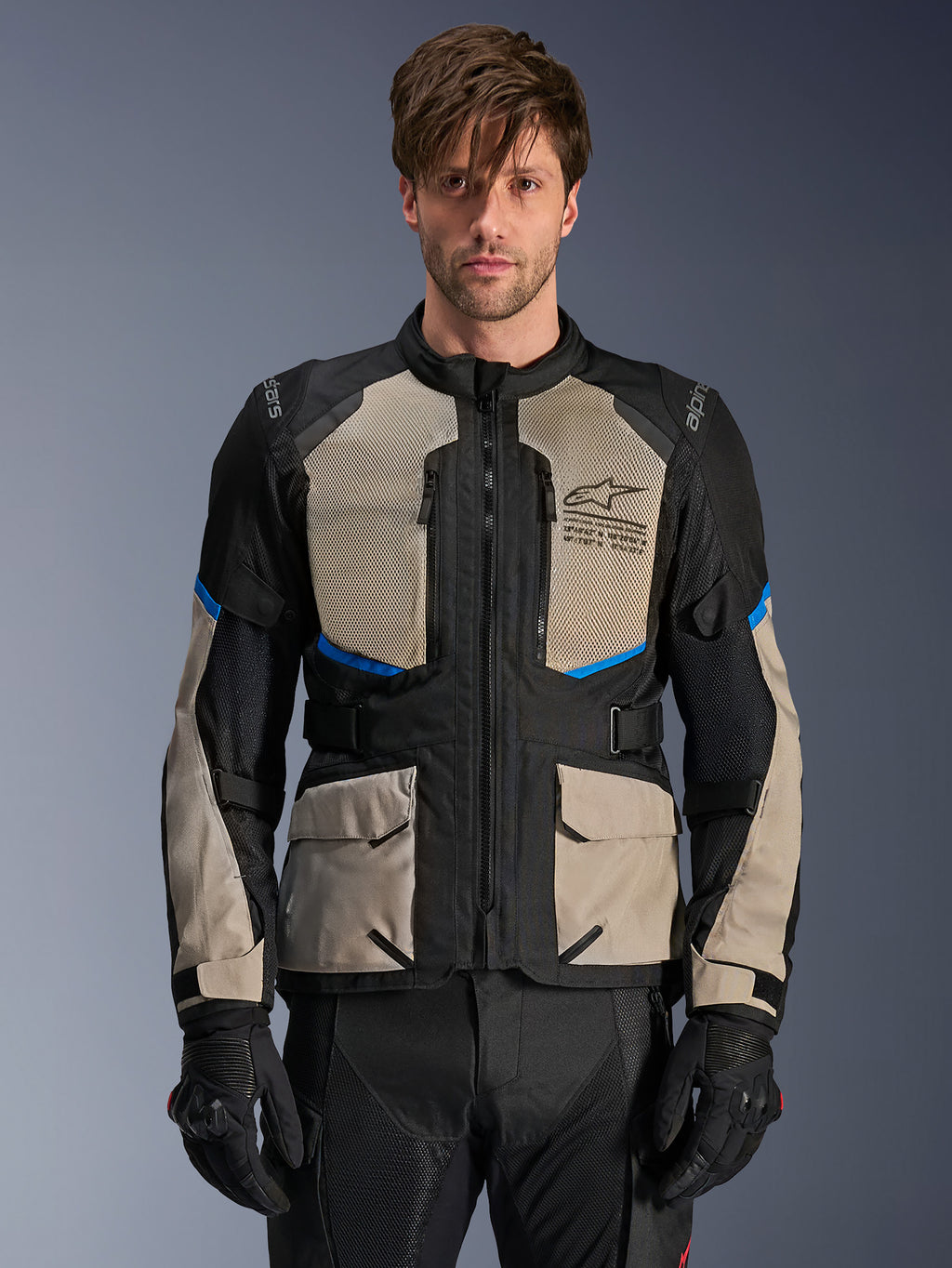 Giacca Alpinestars Andes Air Drystar, giacca adventure touring, nero, grigio laurel e blu brillante, indossata da un modello con guanti neri, dotata di ampi pannelli in rete sul petto per la ventilazione, tasche cargo anteriori e dettagli blu, abbigliamento protettivo da moto touring