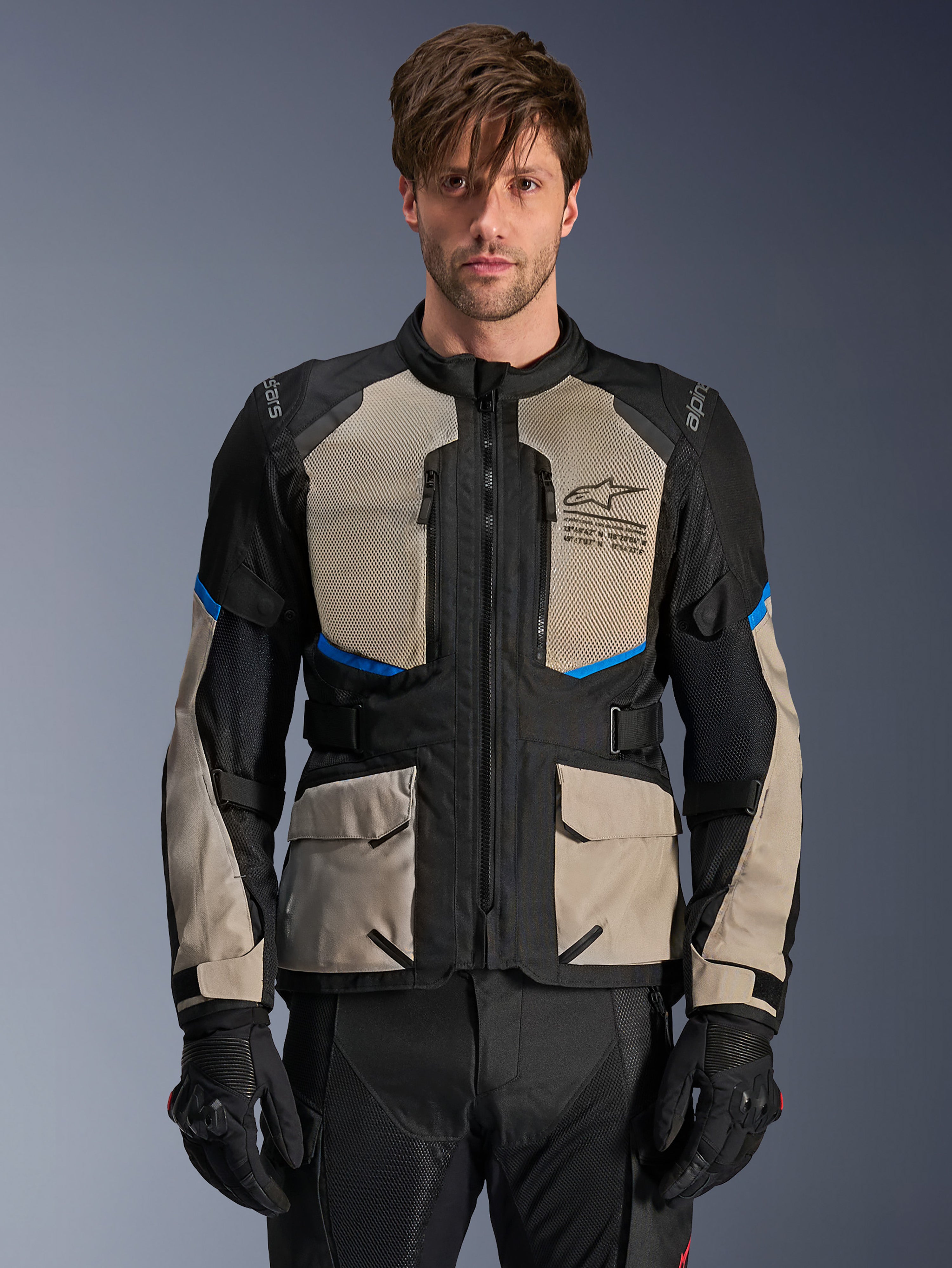 Giacca Alpinestars Andes Air Drystar, giacca adventure touring, nero, grigio laurel e blu brillante, indossata da un modello con guanti neri, dotata di ampi pannelli in rete sul petto per la ventilazione, tasche cargo anteriori e dettagli blu, abbigliamento protettivo da moto touring