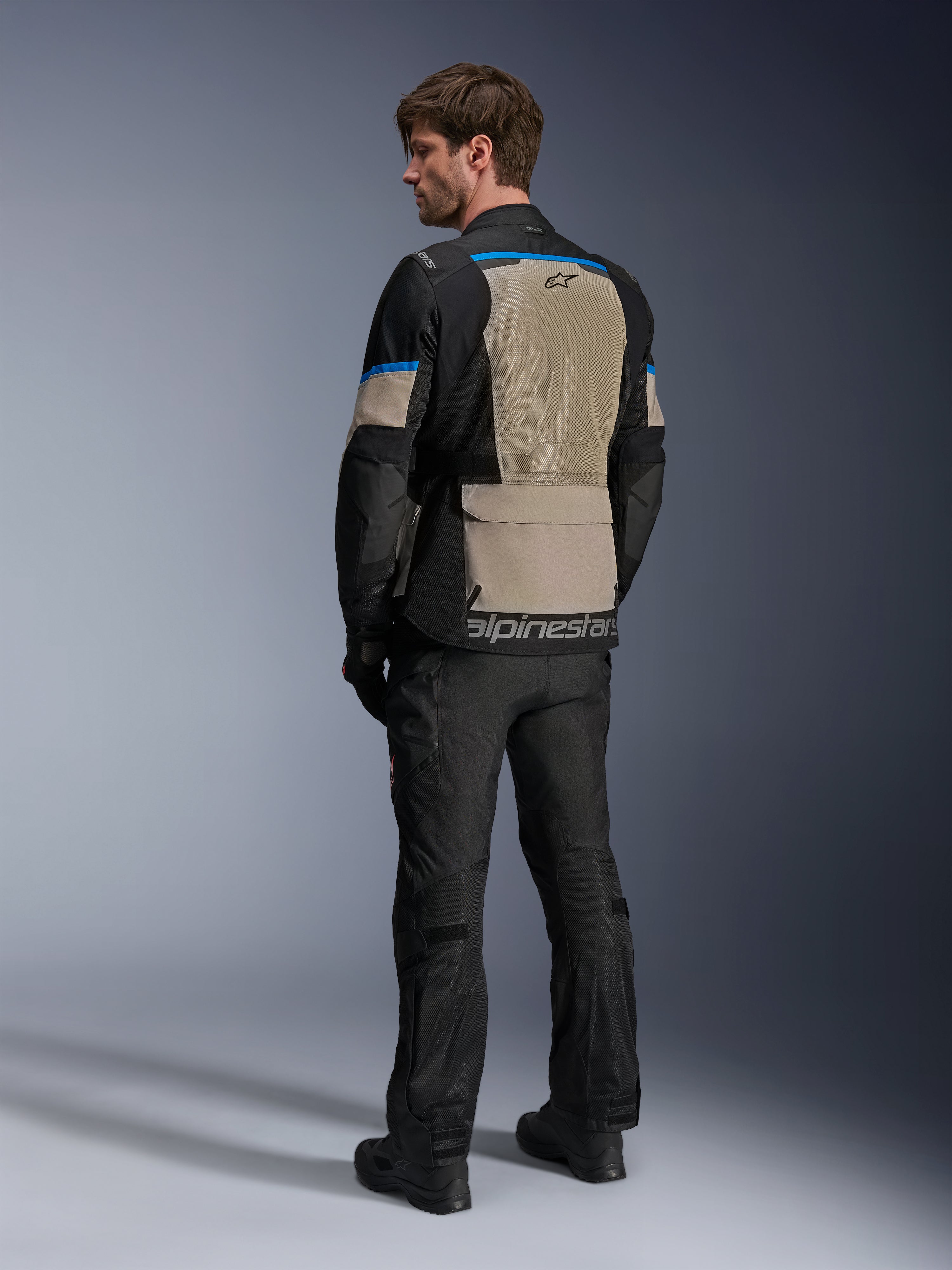 Giacca Alpinestars Andes Air Drystar®, nero, grigio Laurel e blu acceso, vista posteriore che mostra l'ampio pannello di ventilazione in rete sulla schiena, logo sul bordo inferiore, indossata con pantaloni da moto e stivali neri.