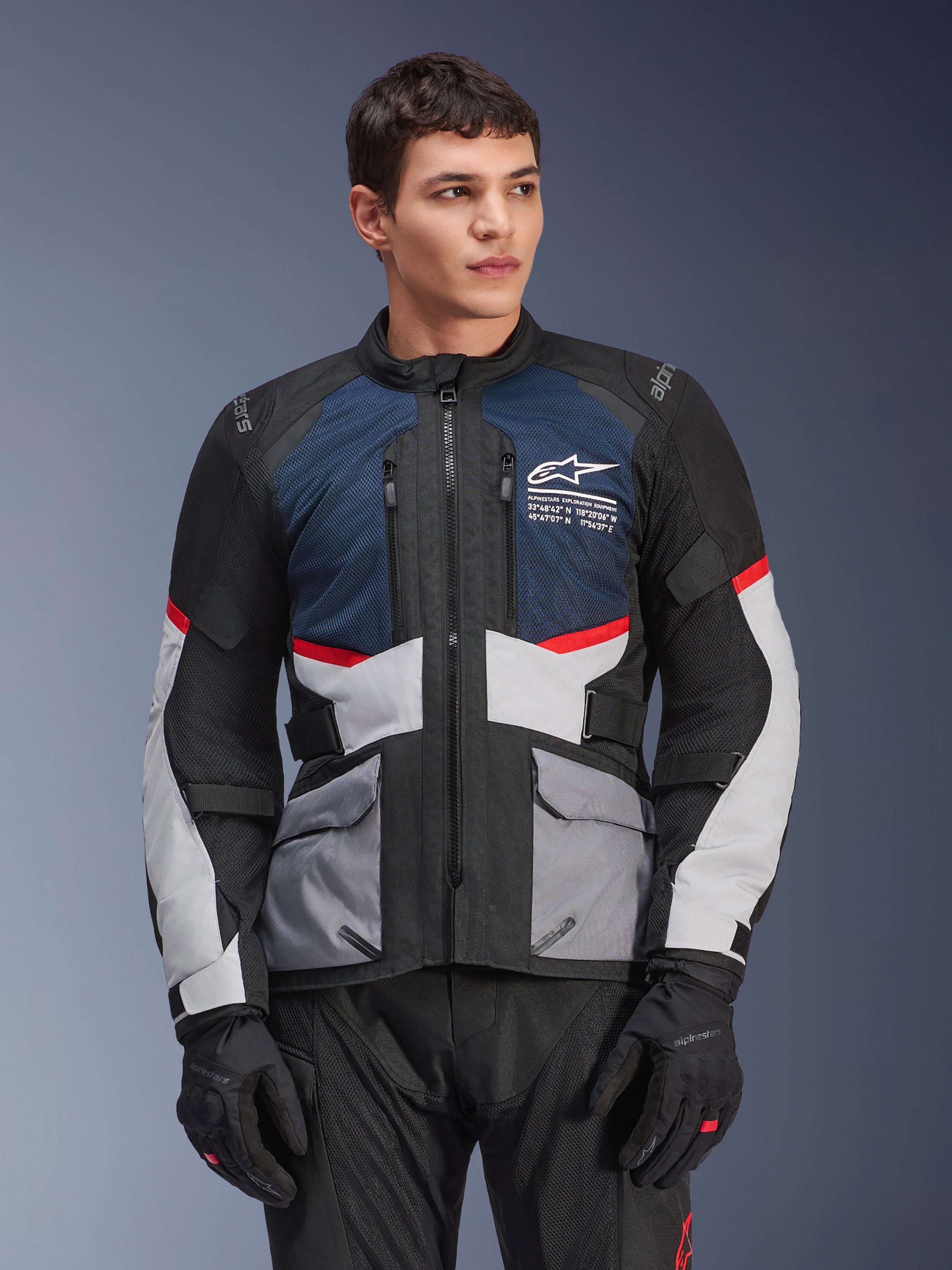 Giacca Alpinestars Andes Air Drystar®, giacca da moto touring, blu scuro, nero e grigio ghiaccio con dettagli rossi, indossata da un modello con guanti neri, dotata di pannelli di ventilazione in rete e tasche utility anteriori, equipaggiamento moderno per avventura e guida su strada