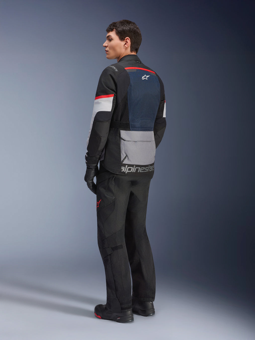 Modello uomo visto di profilo posteriore che indossa la giacca Alpinestars Andes Air Drystar® in Deep Blue Black Ice Gray, abbinata a pantaloni e guanti da moto neri, con pannello posteriore in rete blu, tasca portaoggetti grigia sulla parte bassa della schiena e accenti rossi sulle maniche, abbigliamento professionale per adventure touring.