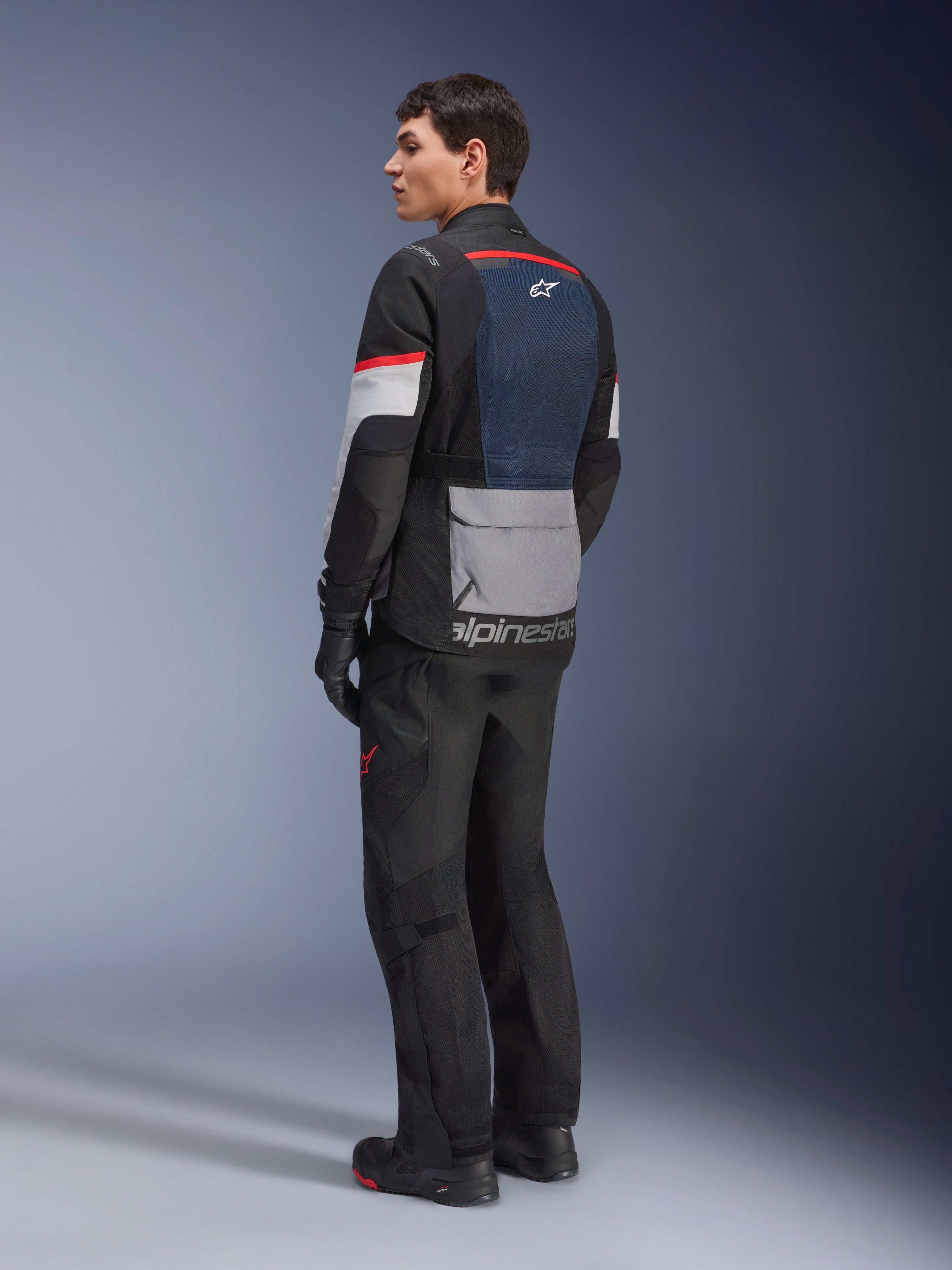 Modello uomo visto di profilo posteriore che indossa la giacca Alpinestars Andes Air Drystar® in Deep Blue Black Ice Gray, abbinata a pantaloni e guanti da moto neri, con pannello posteriore in rete blu, tasca portaoggetti grigia sulla parte bassa della schiena e accenti rossi sulle maniche, abbigliamento professionale per adventure touring.