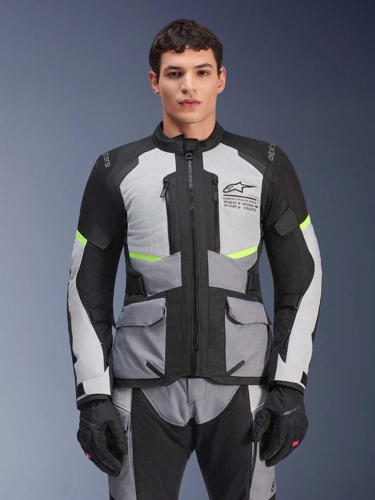 Motociclista che indossa la giacca Alpinestars Andes Air Drystar® in grigio ghiaccio, grigio scuro e nero, con accenti giallo neon e pannelli di ventilazione in rete, abbinata a pantaloni e guanti da moto coordinati, abbigliamento adventure touring