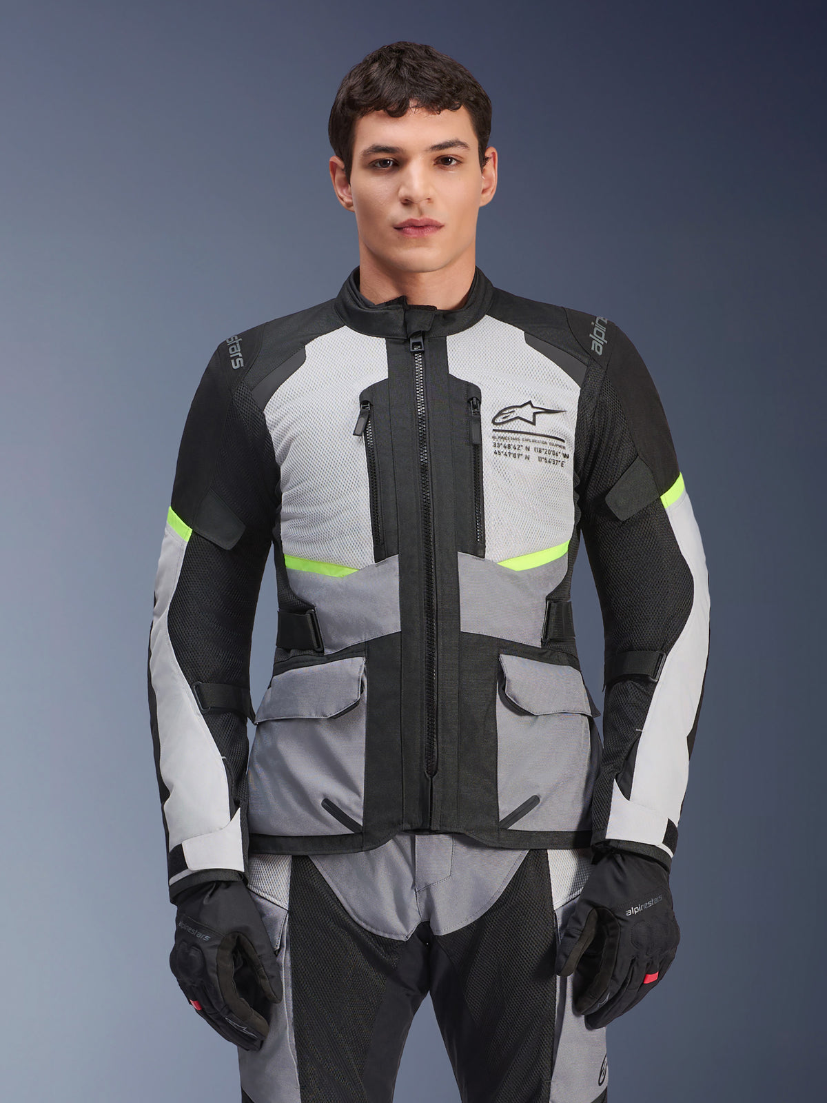 Giacca Alpinestars Andes Air Drystar, giacca da moto adventure touring, grigio ghiaccio e grigio scuro con inserti neri e accenti giallo neon, indossata con pantaloni e guanti coordinati, con pannelli di ventilazione in rete e tasche cargo