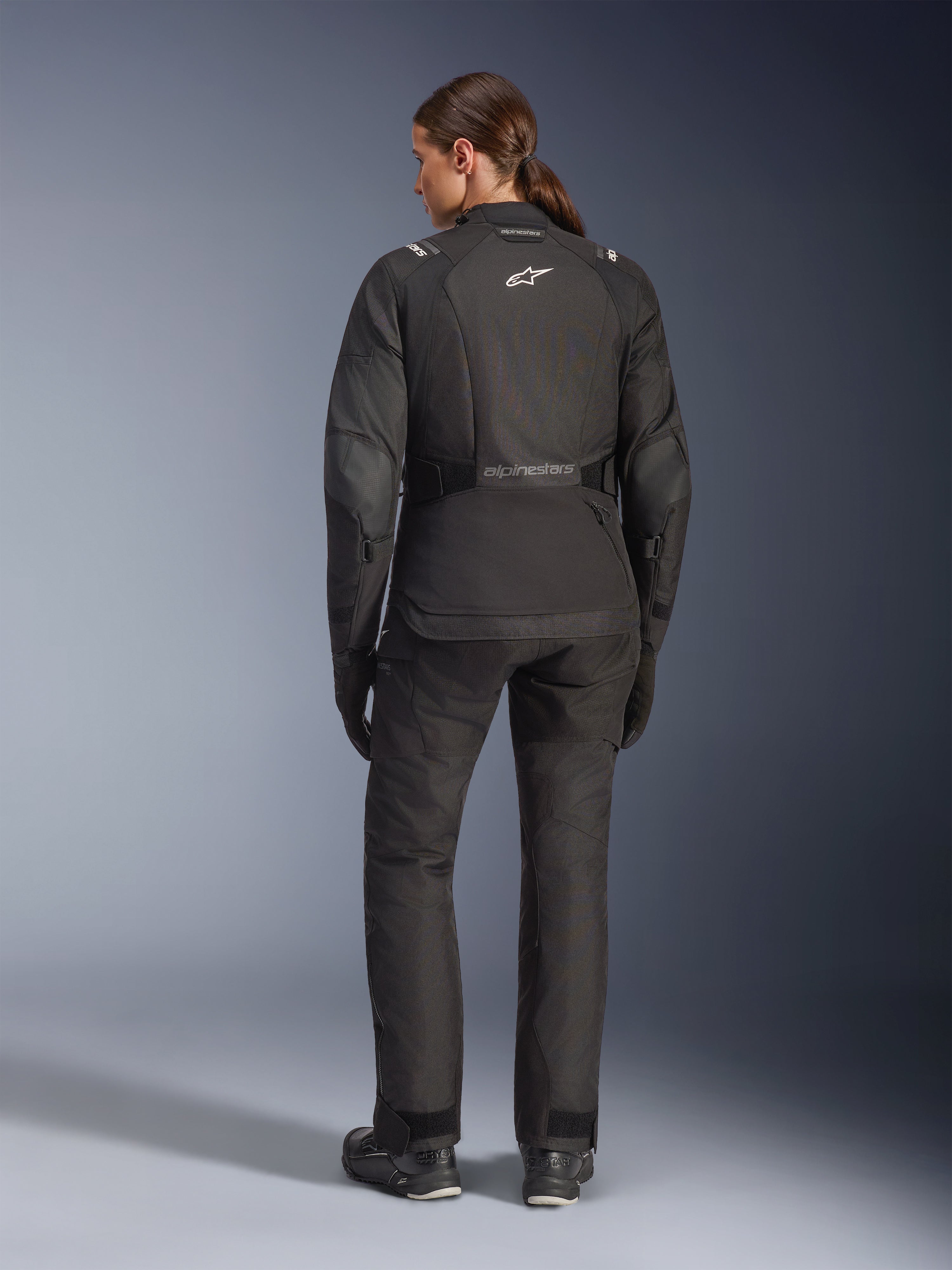 Pilota donna vista da dietro con giacca Alpinestars Stella Andes V4 Drystar®, nera, abbinata a pantaloni da moto coordinati, logo Astar bianco sulla schiena, orlo posteriore allungato, outfit completo da adventure-touring con guanti e stivali neri