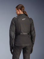 Donna che indossa la giacca Alpinestars Stella Andes V4 Drystar in nero, vista posteriore con logo bianco sulla schiena e pannelli rinforzati sui gomiti, abbinata a pantaloni e guanti neri coordinati, completo da moto adventure touring