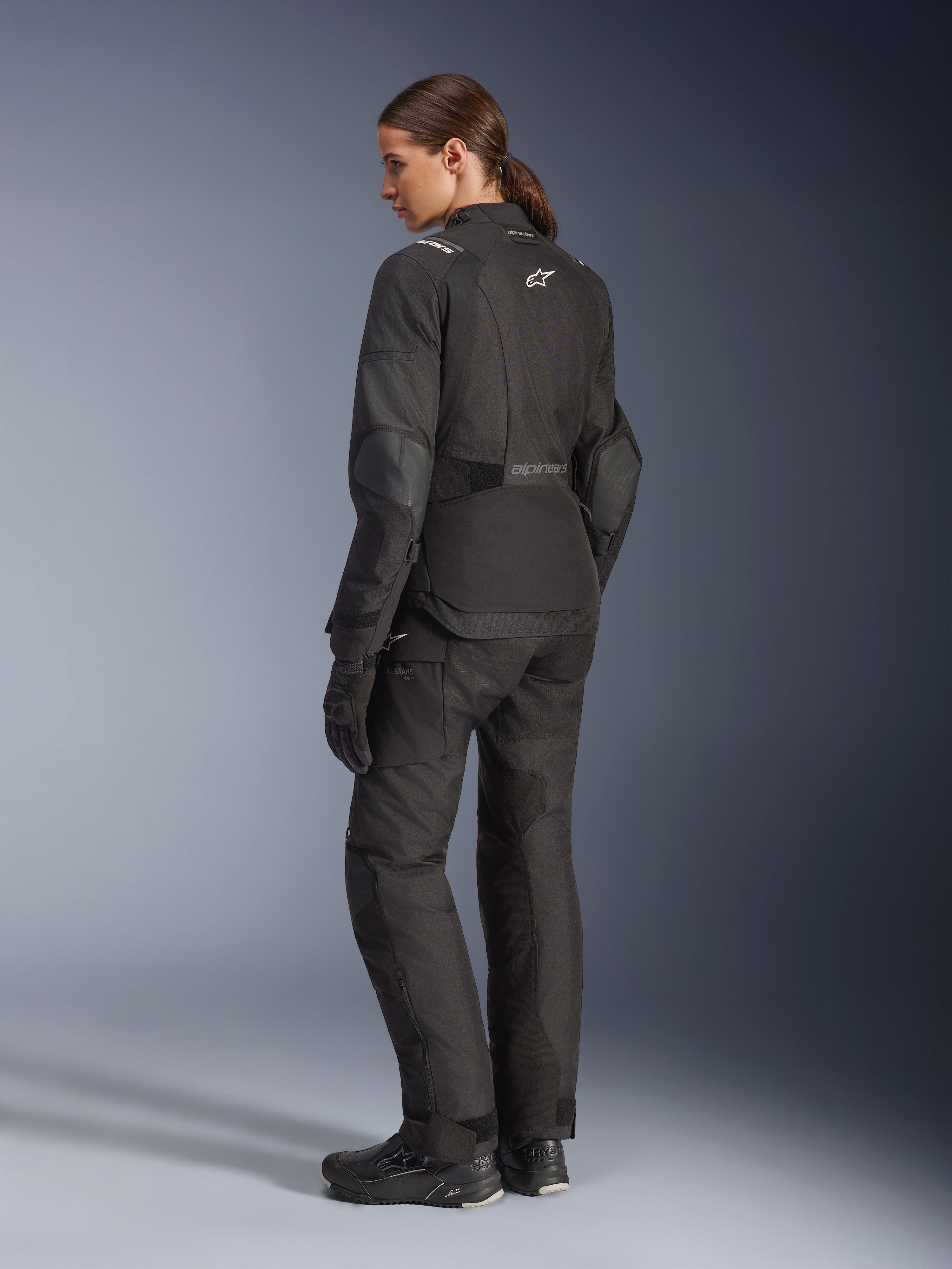 Motociclista donna che indossa la giacca Alpinestars Stella Andes V4 Drystar®, giacca da moto adventure nera, vista posteriore a 3/4, abbinata a pantaloni in tessuto e guanti coordinati, con loghi Alpinestars bianchi sulla schiena e in vita, pannelli protettivi rinforzati e orlo posteriore allungato.