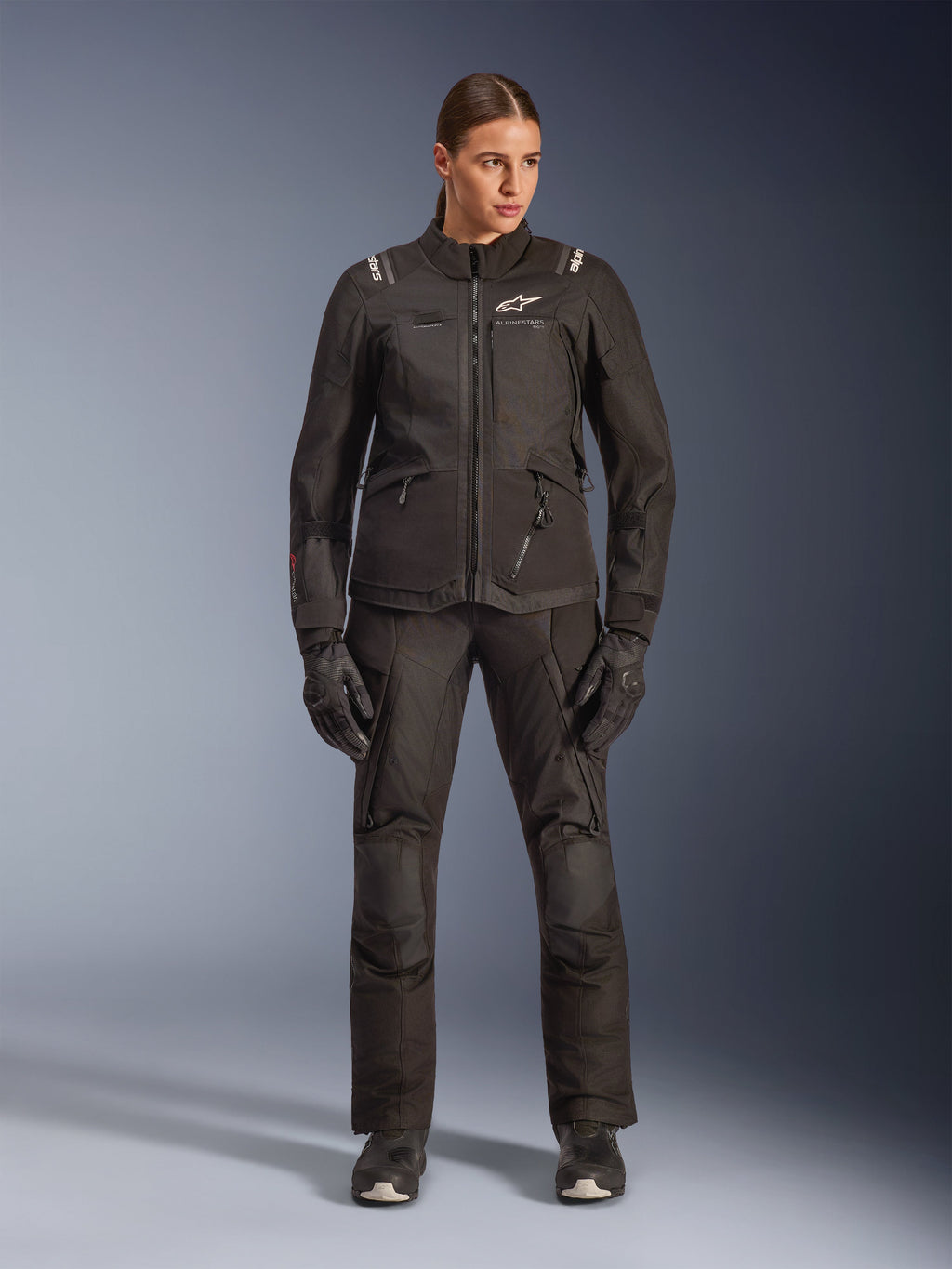 Motociclista donna che indossa la giacca Alpinestars Stella Andes V4 Drystar® in nero, abbinata a pantaloni e guanti adventure-touring coordinati neri, caratterizzata da una costruzione in tessuto tecnico con molteplici tasche multiuso e loghi Alpinestars bianchi, abbigliamento da moto completo per ogni condizione atmosferica