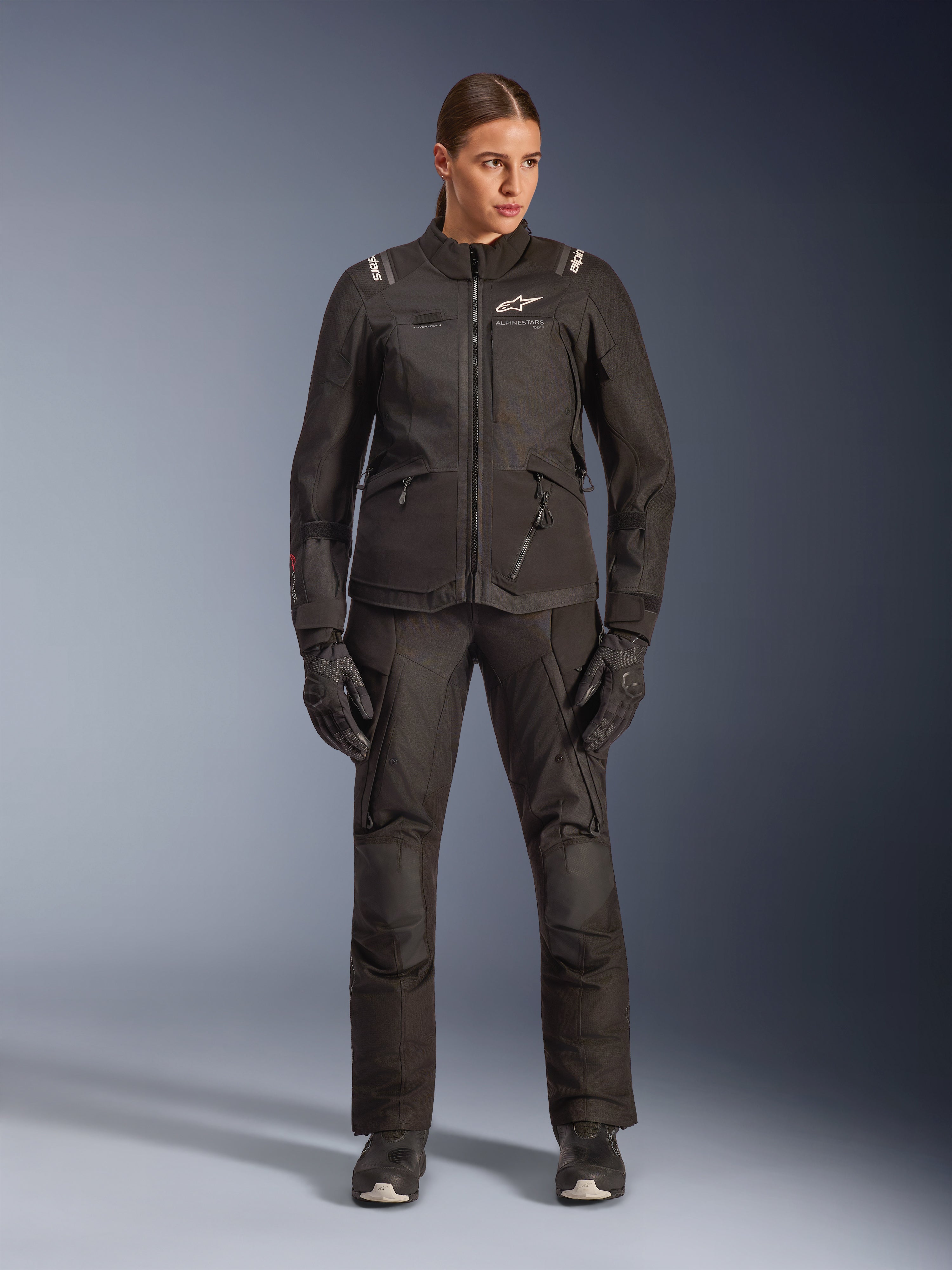 Motociclista donna che indossa la giacca Alpinestars Stella Andes V4 Drystar® in nero, abbinata a pantaloni e guanti adventure-touring coordinati neri, caratterizzata da una costruzione in tessuto tecnico con molteplici tasche multiuso e loghi Alpinestars bianchi, abbigliamento da moto completo per ogni condizione atmosferica