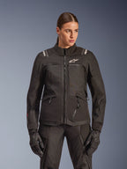 Alpinestars Stella Andes V4 Drystar Jacket, giacca da moto touring da donna, nera, indossata da una modella con pantaloni da moto e guanti coordinati, con chiusura frontale con zip, logo bianco sul petto e branding Alpinestars sulle spalle, abbigliamento tecnico per ogni condizione climatica.
