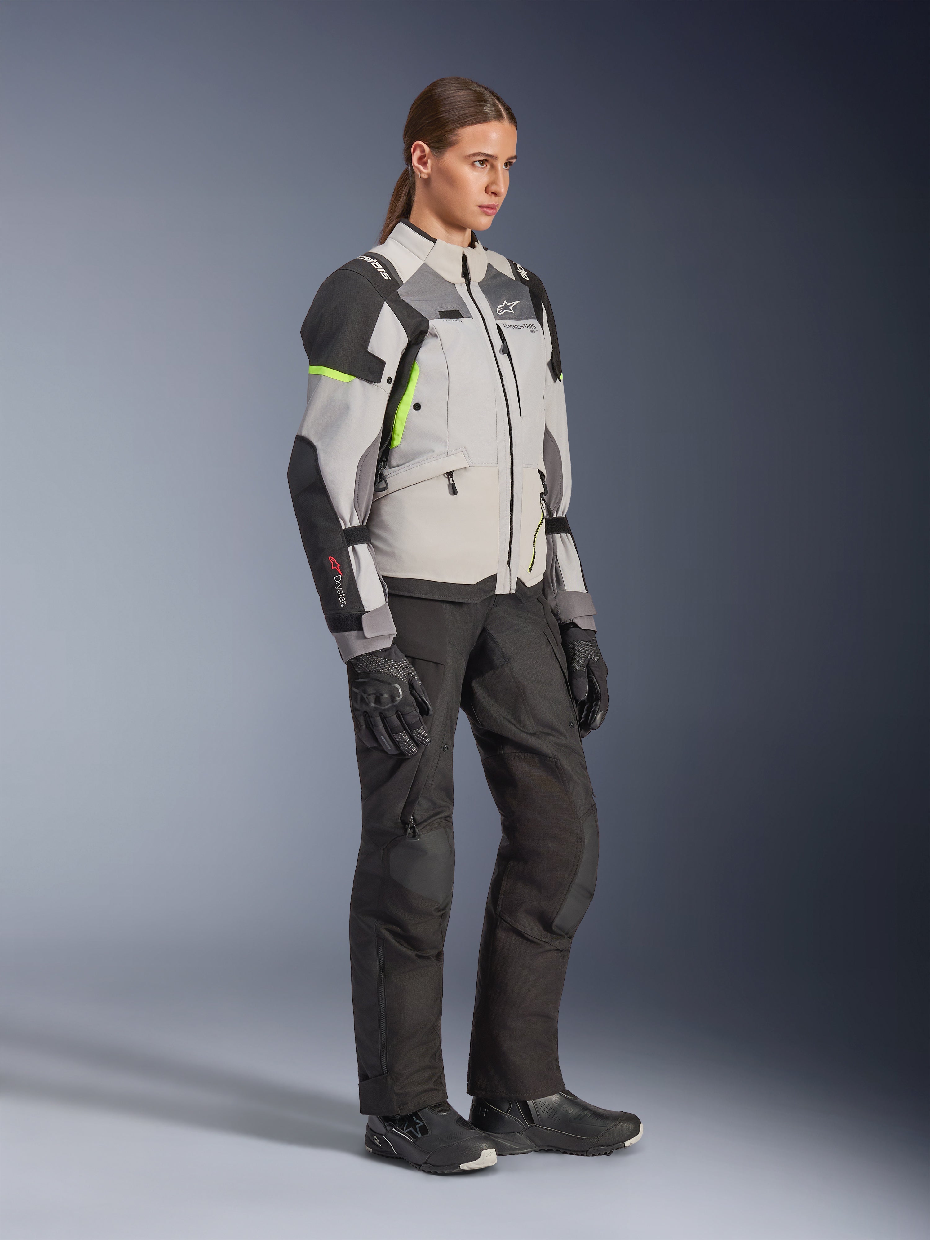 Giacca Alpinestars Stella Andes V4 Drystar® da donna, grigio ghiaccio, grigio scuro e nero con dettagli giallo fluo, indossata da una modella con pantaloni da moto e guanti neri, abbigliamento protettivo da adventure touring per donna