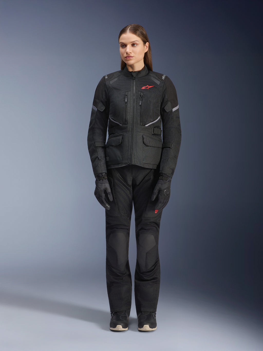 Modella che indossa la giacca Alpinestars Stella Andes Air Drystar® in nero, abbinata a pantaloni da touring e guanti neri coordinati, con ampi pannelli di ventilazione in rete, dettagli del logo in rosso e inserti riflettenti, scatto professionale in studio per abbigliamento da moto da donna.