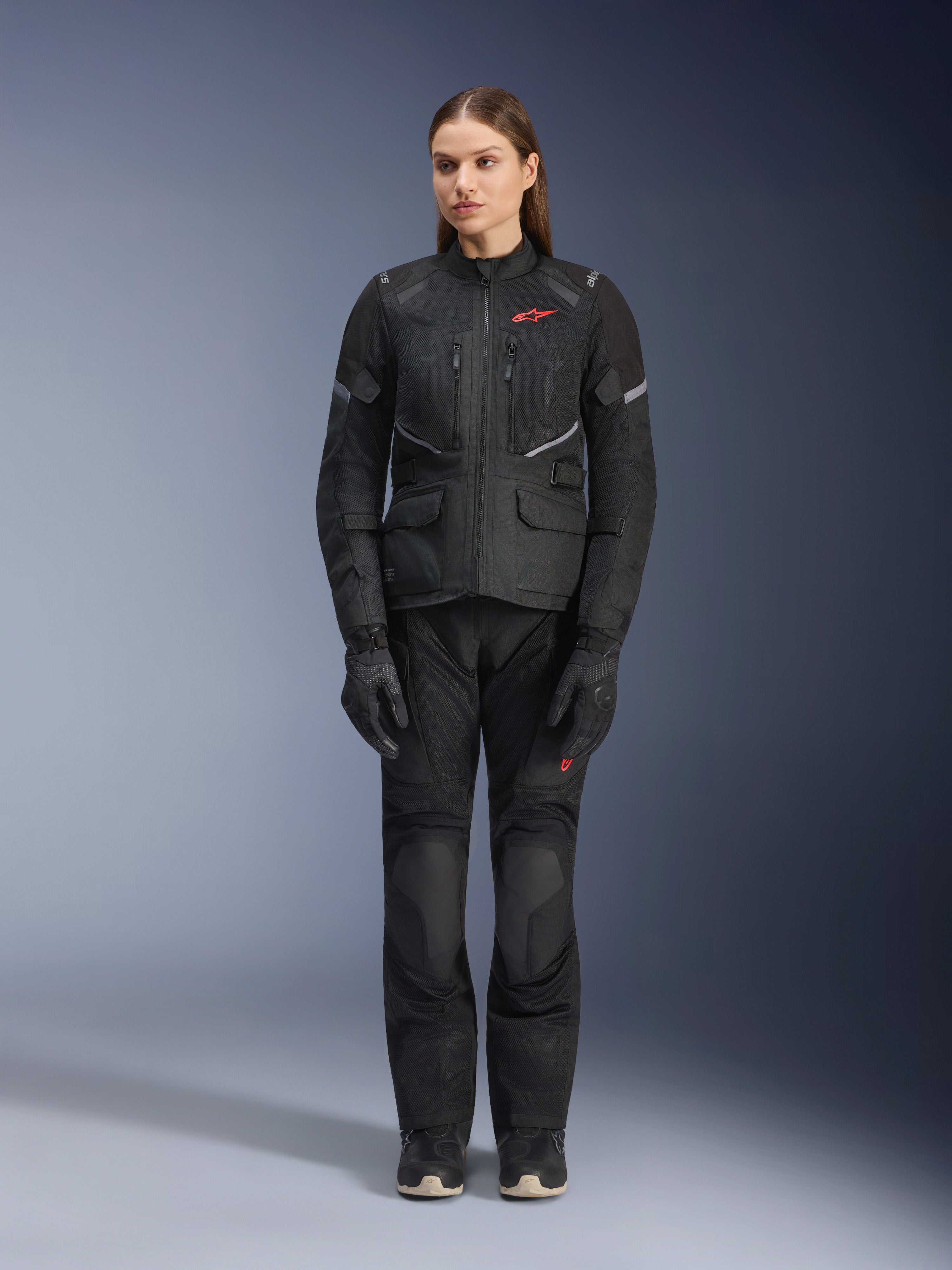 Modella che indossa la giacca Alpinestars Stella Andes Air Drystar® in nero, abbinata a pantaloni da touring e guanti neri coordinati, con ampi pannelli di ventilazione in rete, dettagli del logo in rosso e inserti riflettenti, scatto professionale in studio per abbigliamento da moto da donna.