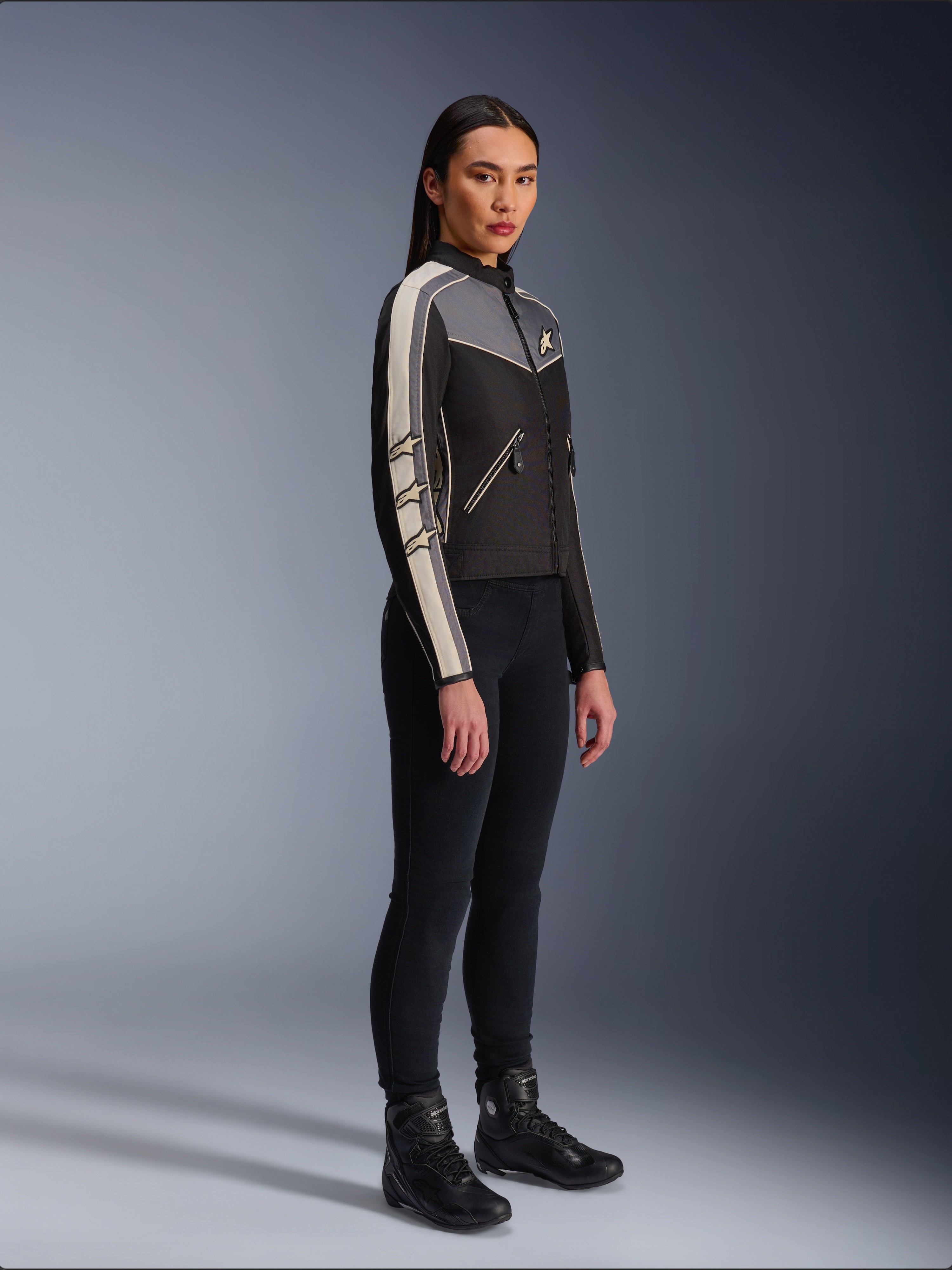 Giacca Alpinestars Stella T-Dyno WR, giacca da moto in tessuto da donna, nero, grigio scuro ed ecru, design d'ispirazione vintage con strisce sulle maniche color panna e loghi a stella, indossata con pantaloni neri e stivali da moto, abbigliamento urban riding d'ispirazione heritage