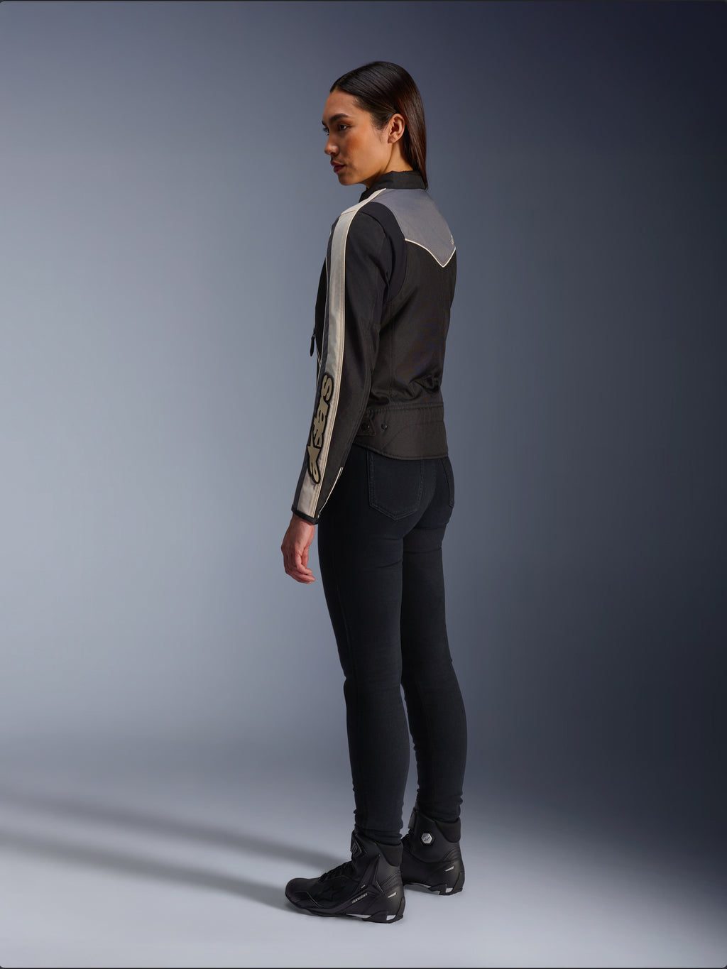 Modella vista di profilo e da dietro che indossa la giacca Alpinestars Stella T-Dyno WR in Nero Grigio Scuro Ecru, caratterizzata da un design di ispirazione vintage con strisce sulle maniche color ecru e un inserto posteriore grigio, abbinata a pantaloni neri e scarpe da moto nere.