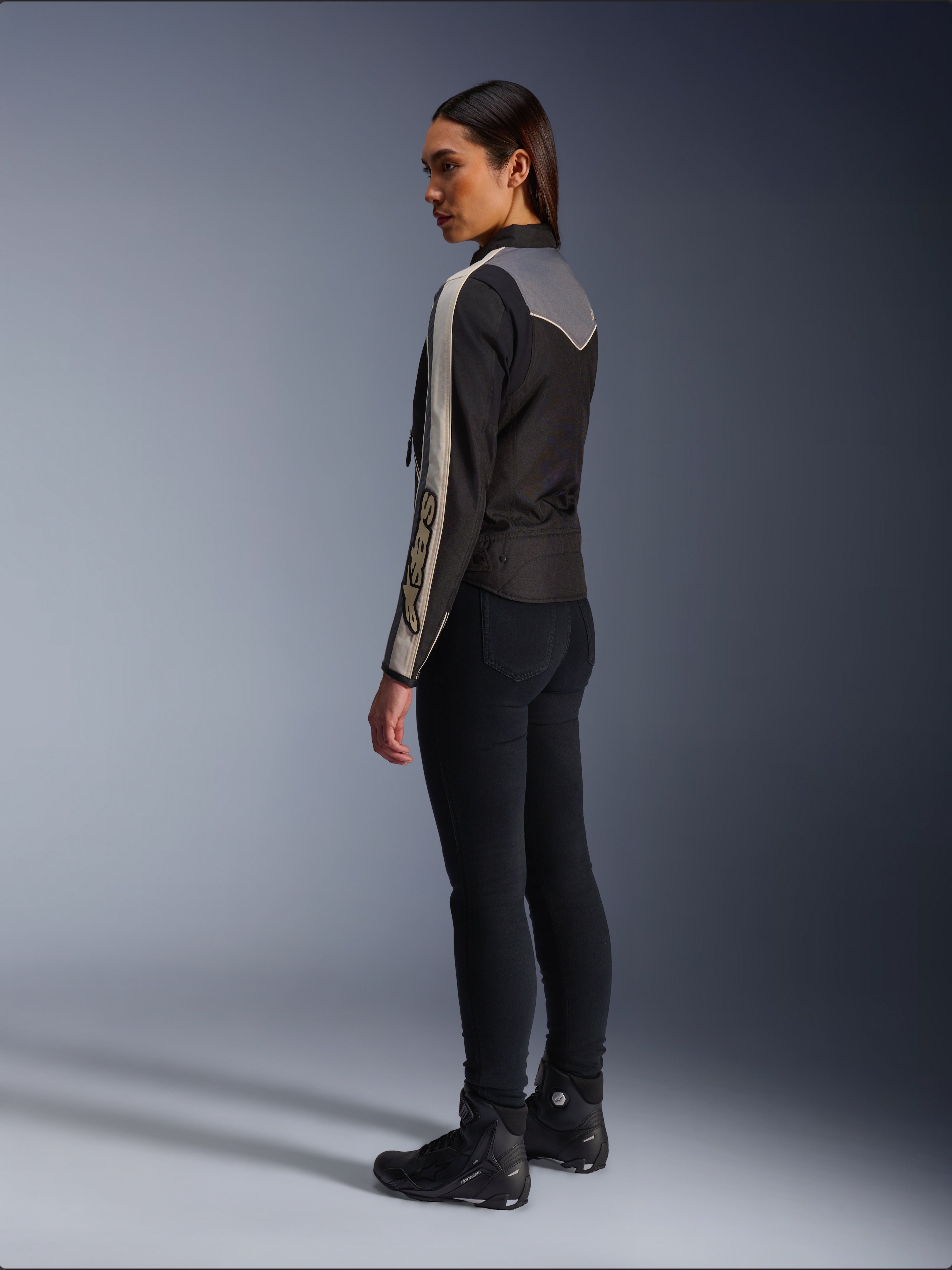 Modella vista di profilo e da dietro che indossa la giacca Alpinestars Stella T-Dyno WR in Nero Grigio Scuro Ecru, caratterizzata da un design di ispirazione vintage con strisce sulle maniche color ecru e un inserto posteriore grigio, abbinata a pantaloni neri e scarpe da moto nere.