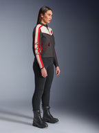Modella in piedi di profilo che indossa la giacca Alpinestars Stella T-Dyno WR in nero, ecru e rosso acceso, con strisce racing sulle maniche e loghi a stella, abbinata a pantaloni neri e stivali stringati per uno stile motociclistico urbano.