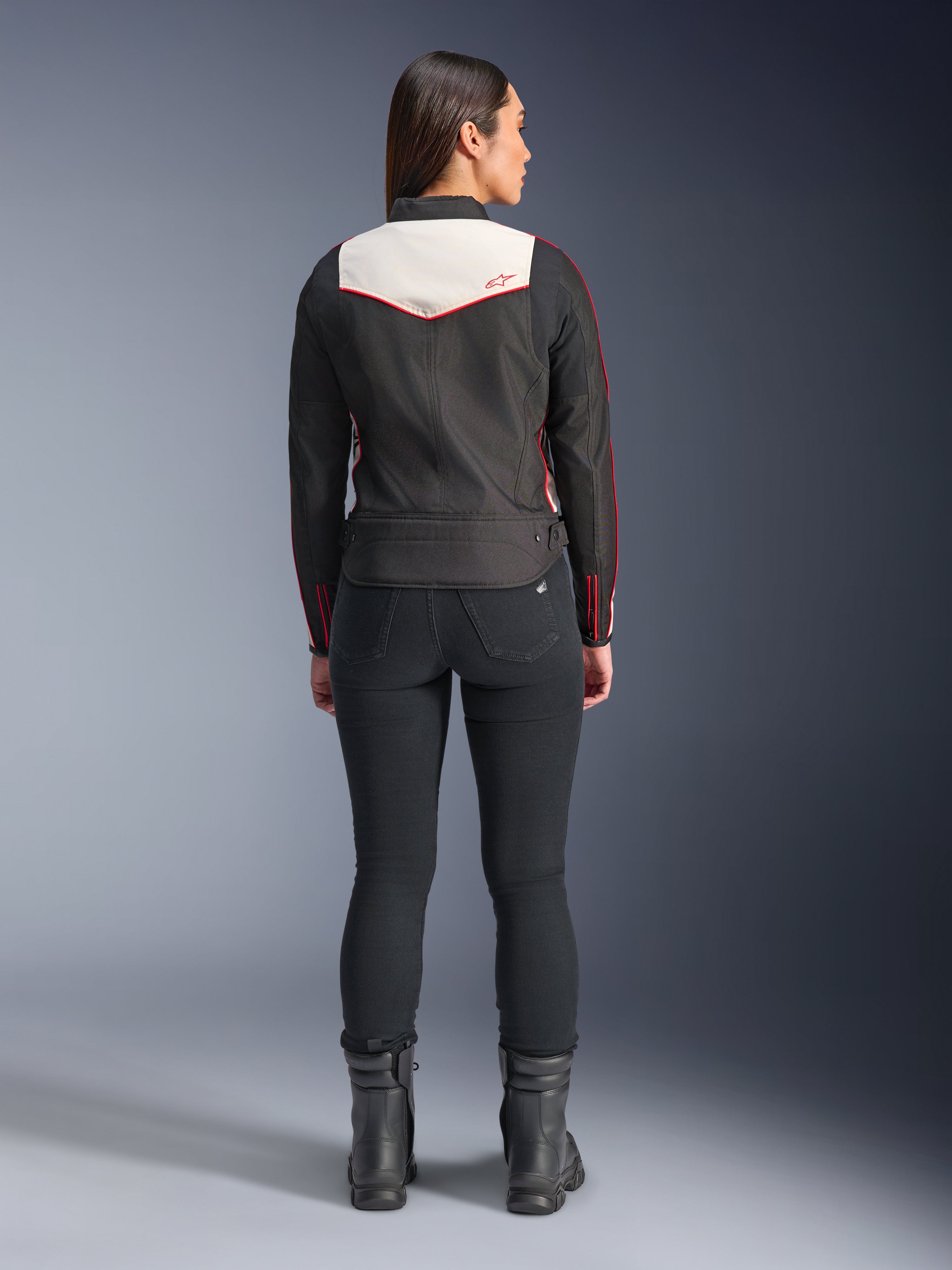 Vista posteriore di una modella che indossa la giacca Alpinestars Stella T-Dyno WR in Nero Ecru Rosso Brillante, caratterizzata da un evidente pannello posteriore color crema e dettagli del logo rossi, abbinata a pantaloni e stivali neri, una giacca da moto in tessuto d'ispirazione vintage.
