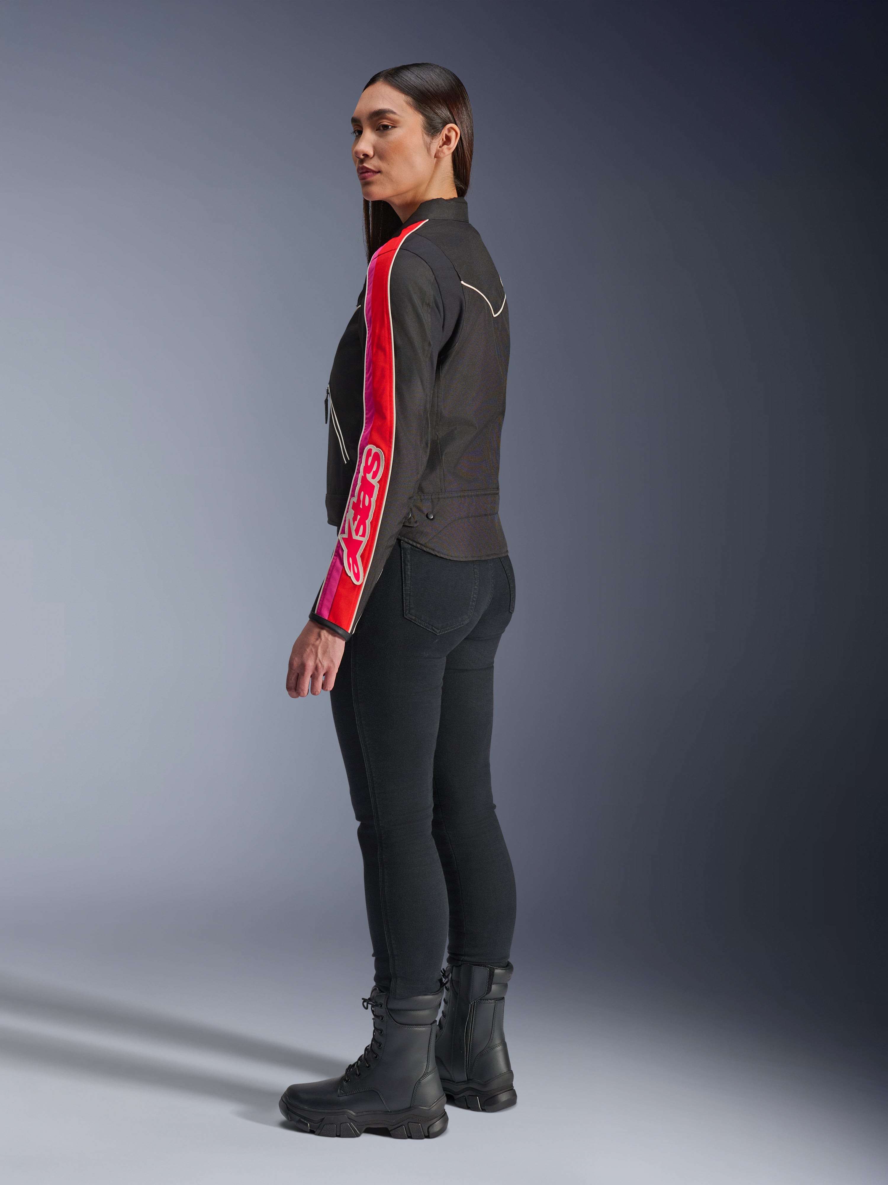 Modella che indossa la giacca Alpinestars Stella T-Dyno WR in Nero Rosso Brillante Fucsia, caratterizzata da audaci strisce rosse e rosa con il marchio Alpinestars sulla manica, abbinata a pantaloni da guida neri e stivali da moto neri in un set in studio.