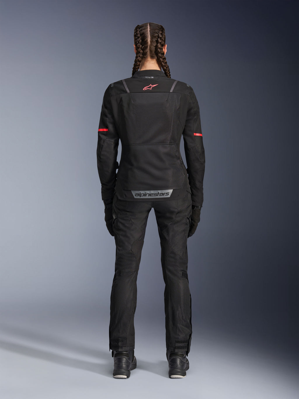 Giacca Alpinestars Stella ST-2 Air, giacca da moto da donna, nero, vista posteriore con logo rosso sulla parte superiore della schiena e marchio grigio sulla parte inferiore della schiena, indossata con pantaloni e stivali da moto neri coordinati, design touring moderno con pannelli in mesh e dettagli riflettenti