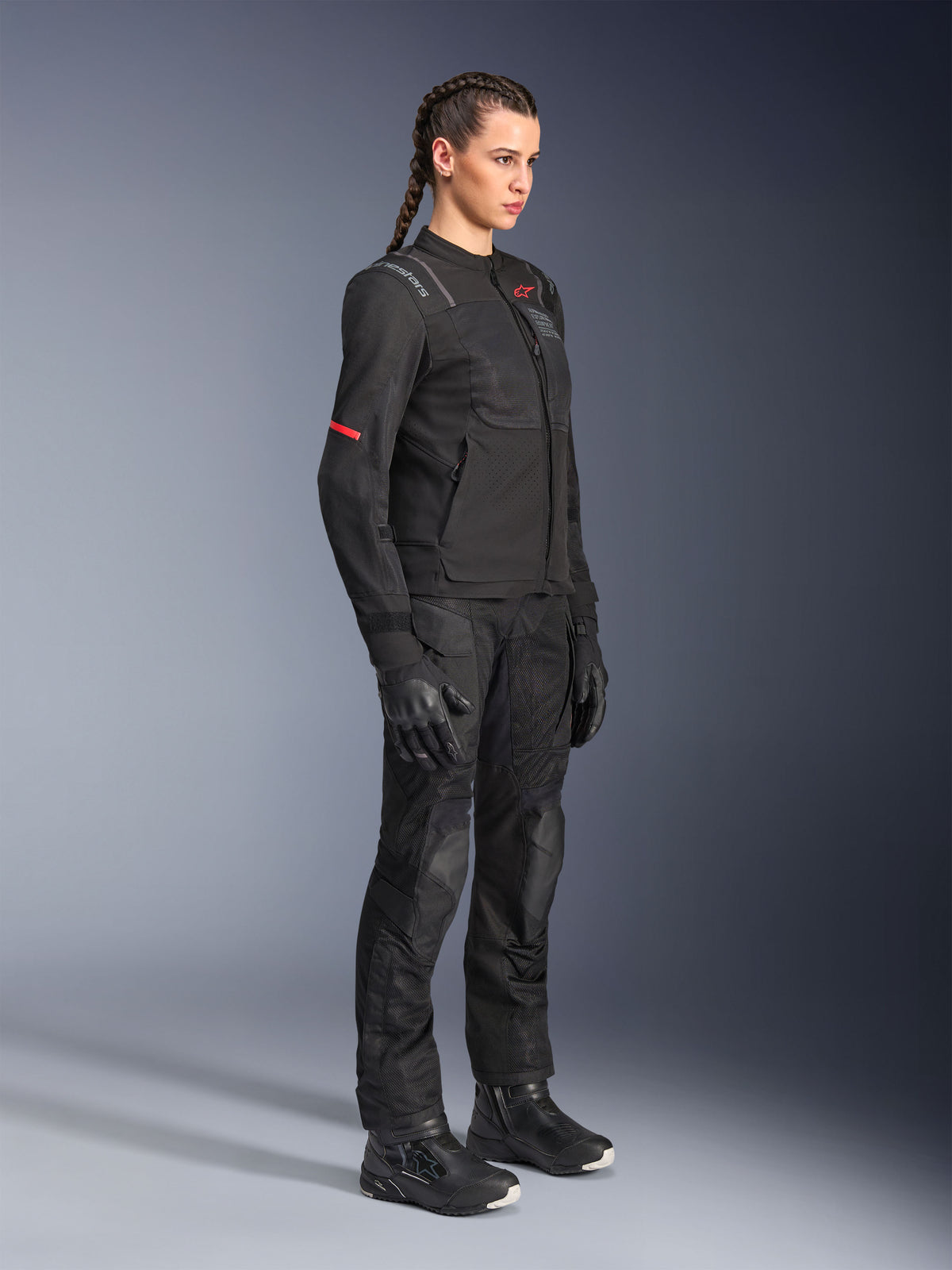 Modella che indossa la giacca Alpinestars Stella ST-2 Air in nero, abbinata a pantaloni da moto, guanti e stivali neri coordinati, con pannelli di ventilazione in mesh e discreti dettagli rossi, outfit da touring moderno ed elegante