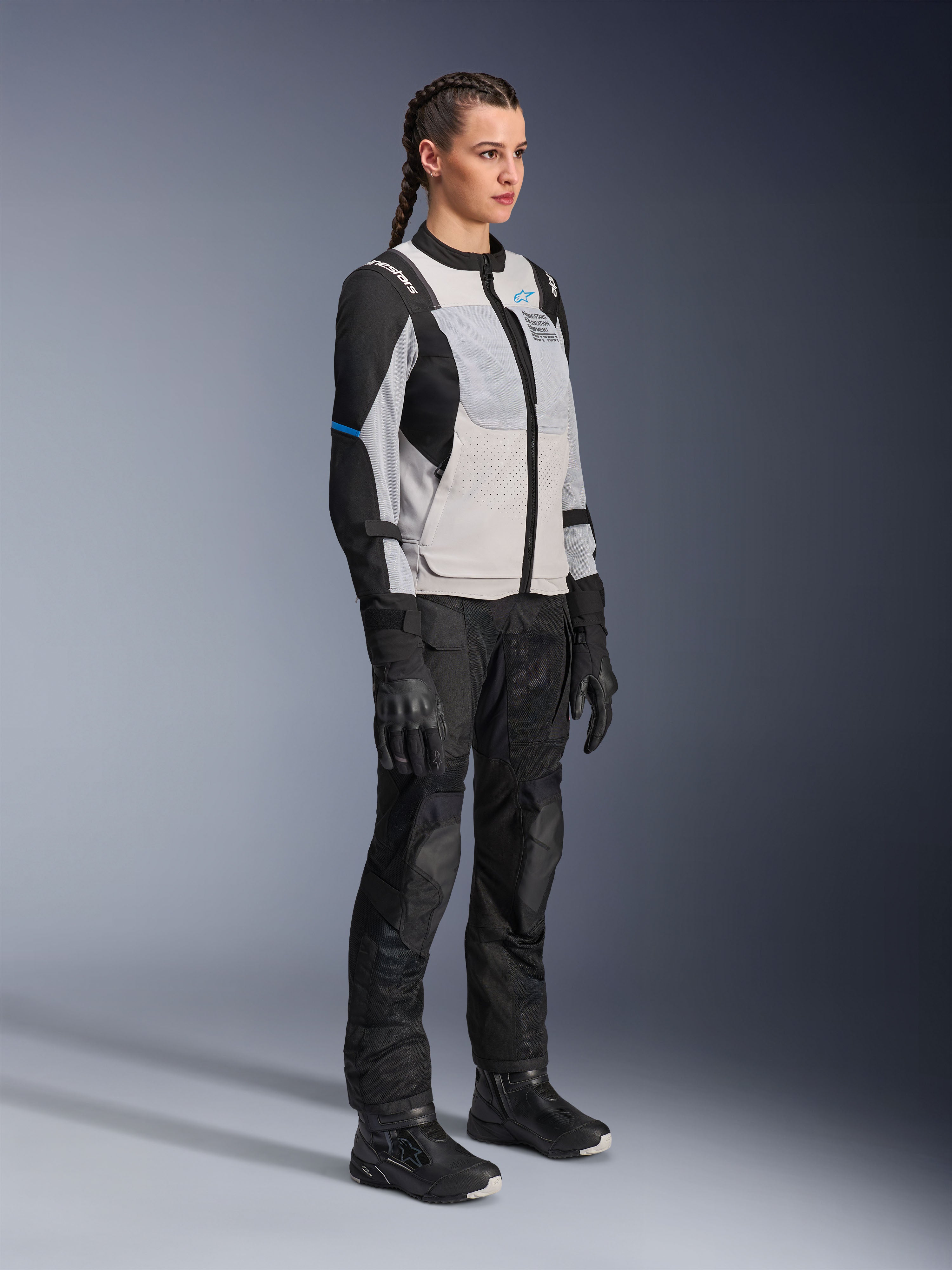 Modella che indossa la giacca Alpinestars Stella ST-2 Air in Grigio Ghiaccio e Nero, giacca tecnica da moto da donna con pannelli in rete e costruzione softshell, abbinata a pantaloni e stivali da touring adventure neri, caratterizzata da accenti blu e un design da guida moderno