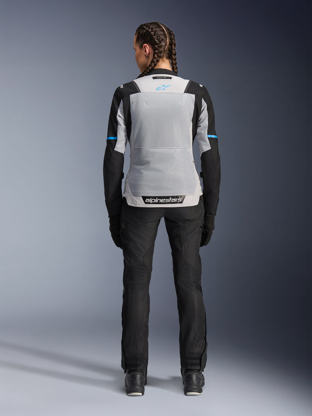 Alpinestars Stella ST-2 Air Jacket, Ice Gray Black, vista posteriore su modella, con ampio pannello posteriore in rete grigio chiaro, maniche nere con strisce blu, logo Alpinestars sulla parte bassa della schiena, abbinata a pantaloni tecnici e stivali neri, abbigliamento touring estivo
