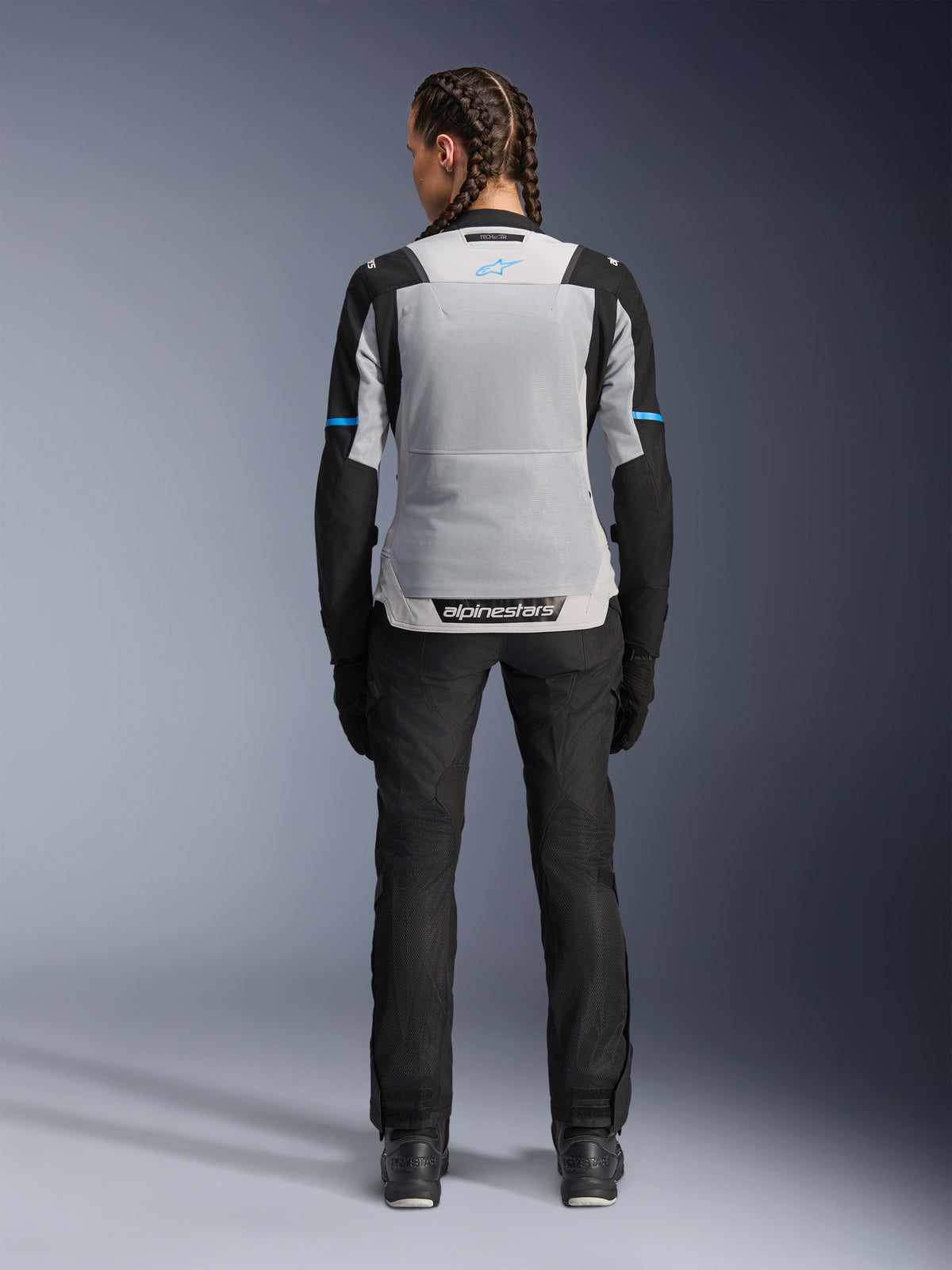Alpinestars Stella ST-2 Air Jacket, Ice Gray Black, vista posteriore su modella, con ampio pannello posteriore in rete grigio chiaro, maniche nere con strisce blu, logo Alpinestars sulla parte bassa della schiena, abbinata a pantaloni tecnici e stivali neri, abbigliamento touring estivo
