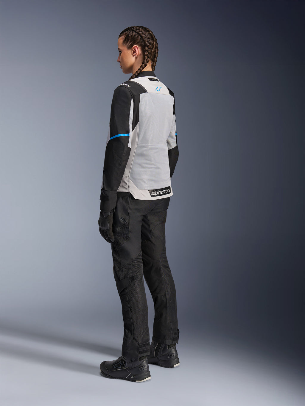 Giacca Alpinestars Stella ST-2 Air in Ice Gray e Nero indossata da una pilota, vista posteriore di tre quarti che evidenzia l'ampio pannello posteriore in mesh grigio chiaro, le maniche nere con dettaglio blu e il logo Alpinestars blu, abbinata a pantaloni, guanti e stivali neri per un outfit completo da adventure touring