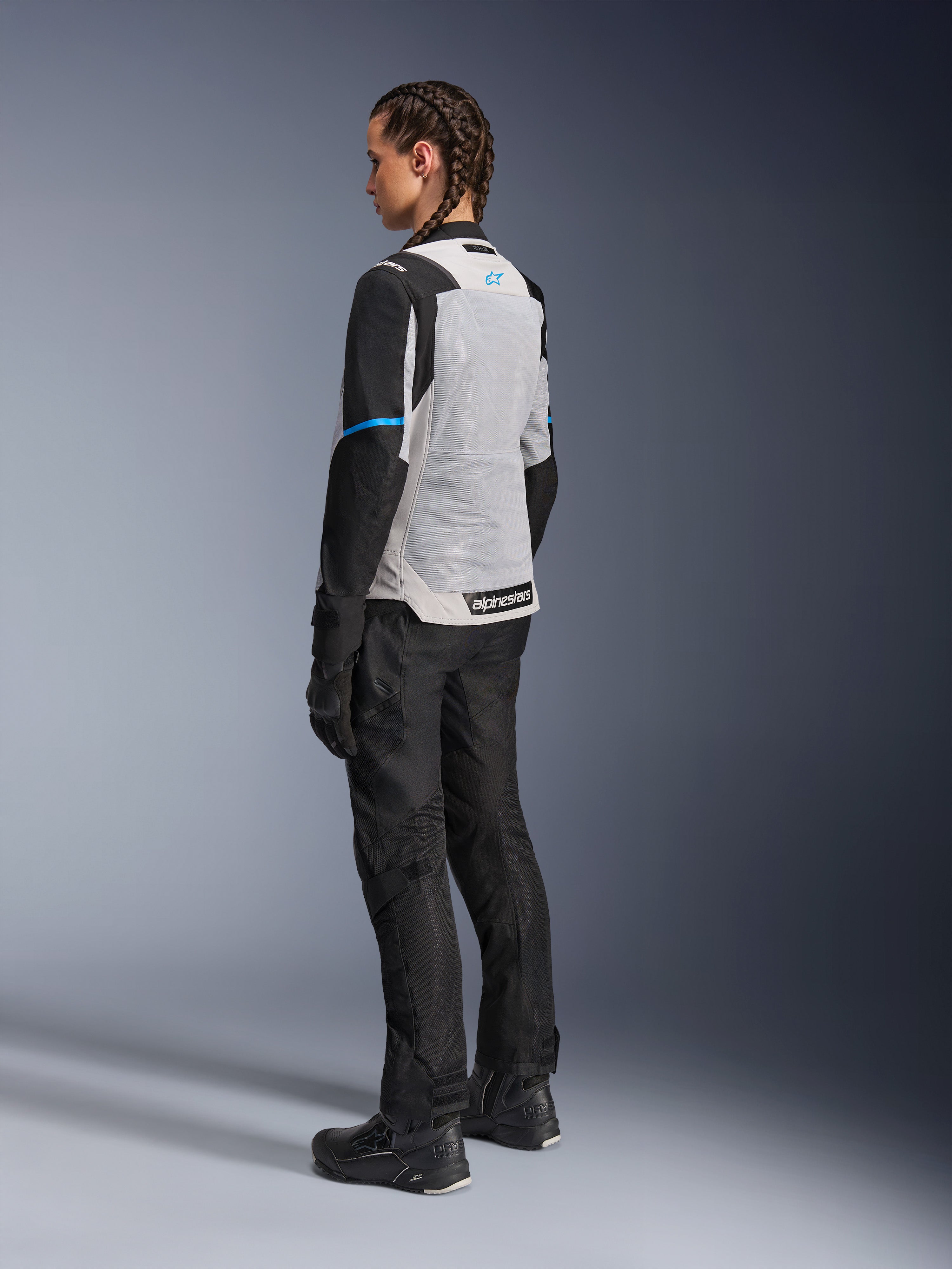 Giacca Alpinestars Stella ST-2 Air in Ice Gray e Nero indossata da una pilota, vista posteriore di tre quarti che evidenzia l'ampio pannello posteriore in mesh grigio chiaro, le maniche nere con dettaglio blu e il logo Alpinestars blu, abbinata a pantaloni, guanti e stivali neri per un outfit completo da adventure touring