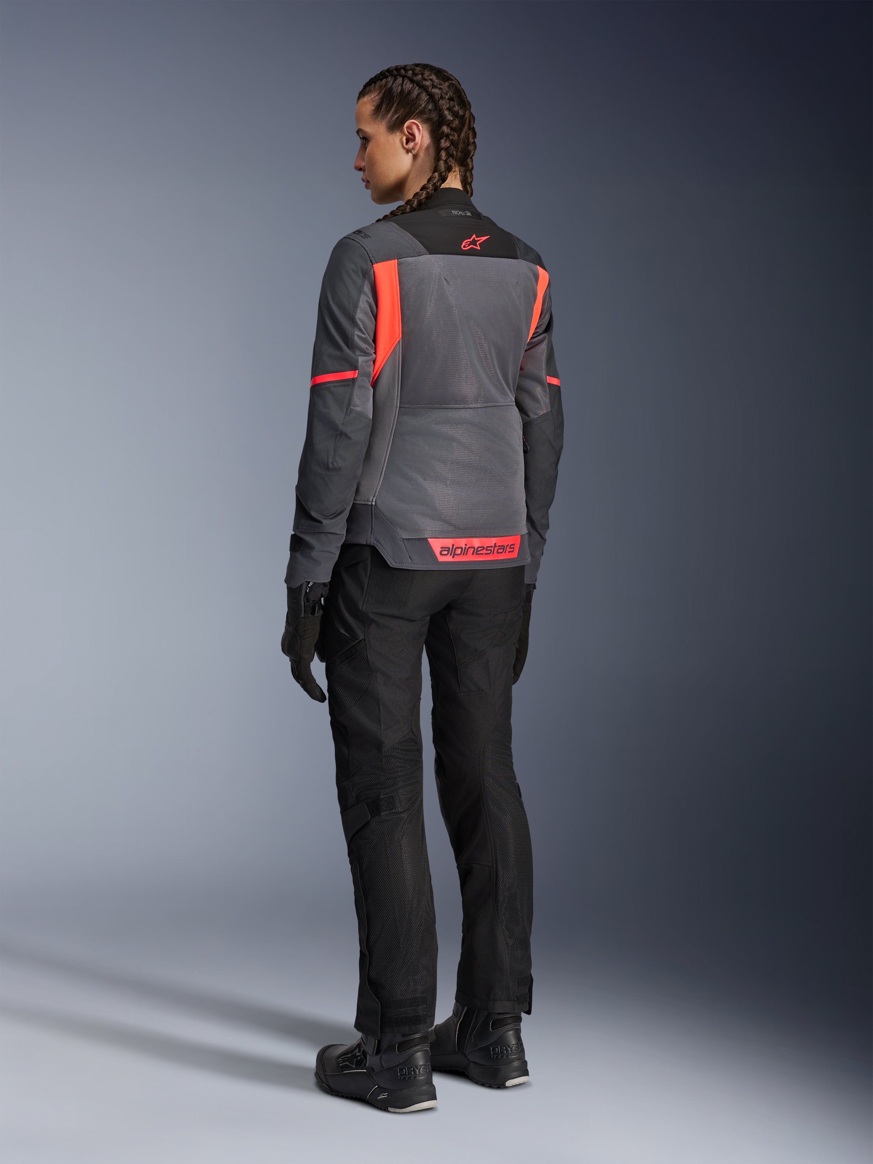 Donna che indossa la giacca Alpinestars Stella St-2 Air, giacca da moto touring, Deep Gray e Dark Gray con accenti Fluo Coral, abbinata a pantaloni da guida e guanti neri, vista posteriore che mostra ampi pannelli in mesh e marchio riflettente corallo, progettata per avventure in moto estive