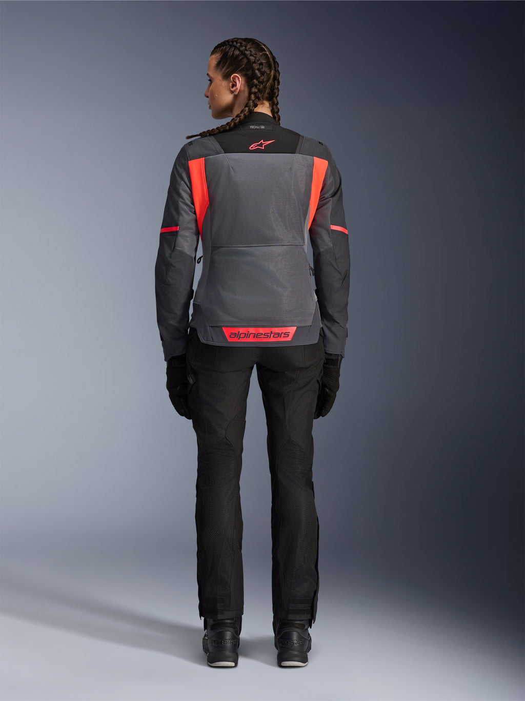 Vista posteriore di una modella che indossa la giacca Alpinestars Stella St-2 Air da donna in Deep Gray e Dark Gray con accenti Fluo Coral, con ampi pannelli di ventilazione in mesh sulla schiena e dettagli del logo riflettenti, abbinata a pantaloni e guanti da moto neri.
