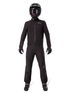 Motociclista che indossa la fodera per pantaloni Alpinestars AMT-8 Stretch Drystar® XF e la giacca coordinata, abbigliamento tecnico completamente nero impermeabile e antivento, con casco, guanti e stivali neri, design elegante per adventure touring.