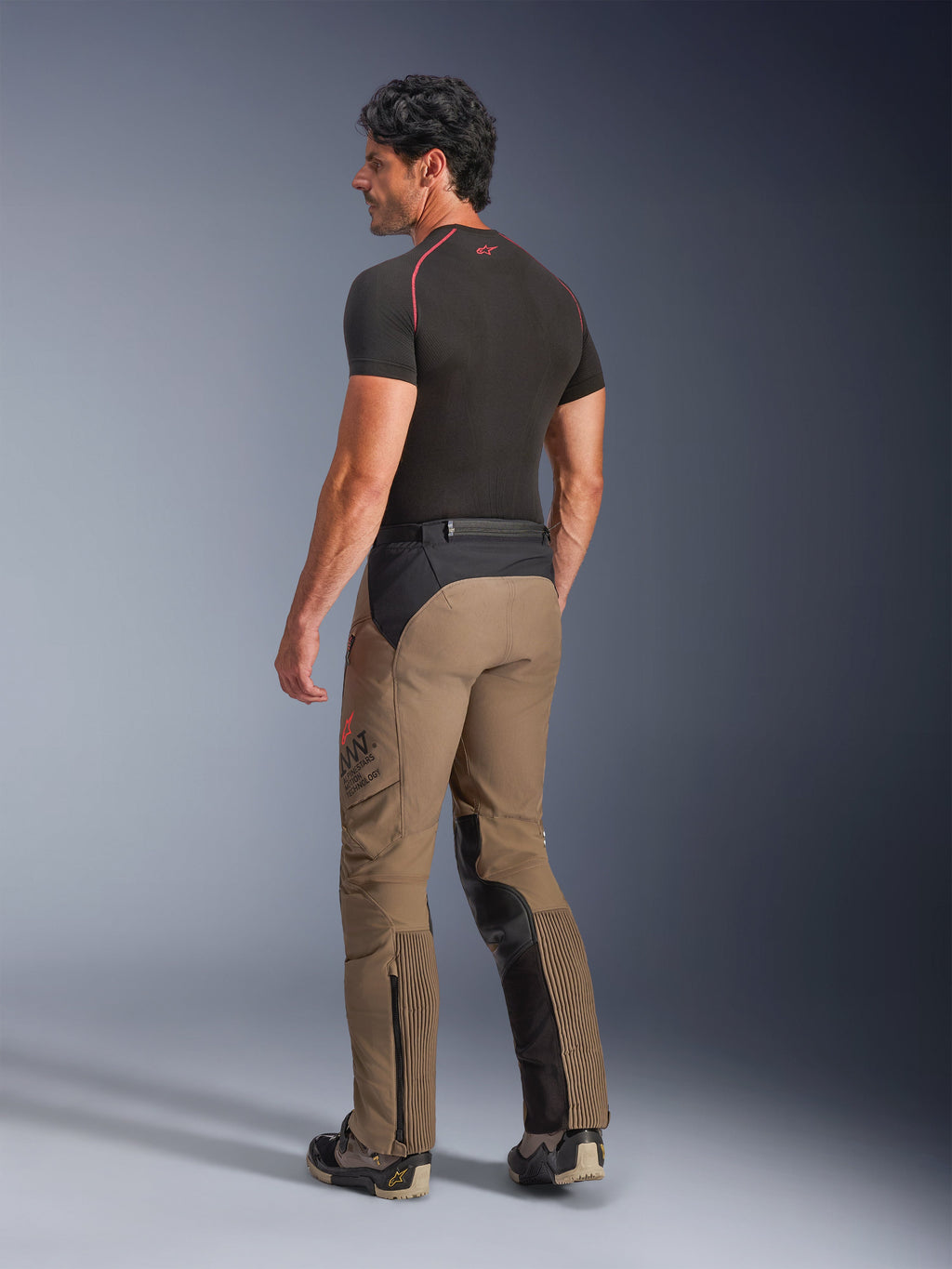 Pantaloni Alpinestars AMT-8 Stretch Drystar® XF in Walnut Black, pantaloni da moto adventure-touring visti da una prospettiva posteriore di tre quarti, in tessuto marrone noce con pannelli neri su seduta e interno gamba, inserti elasticizzati a fisarmonica sui polpacci e marchio AMT laterale, indossati con base layer nero.