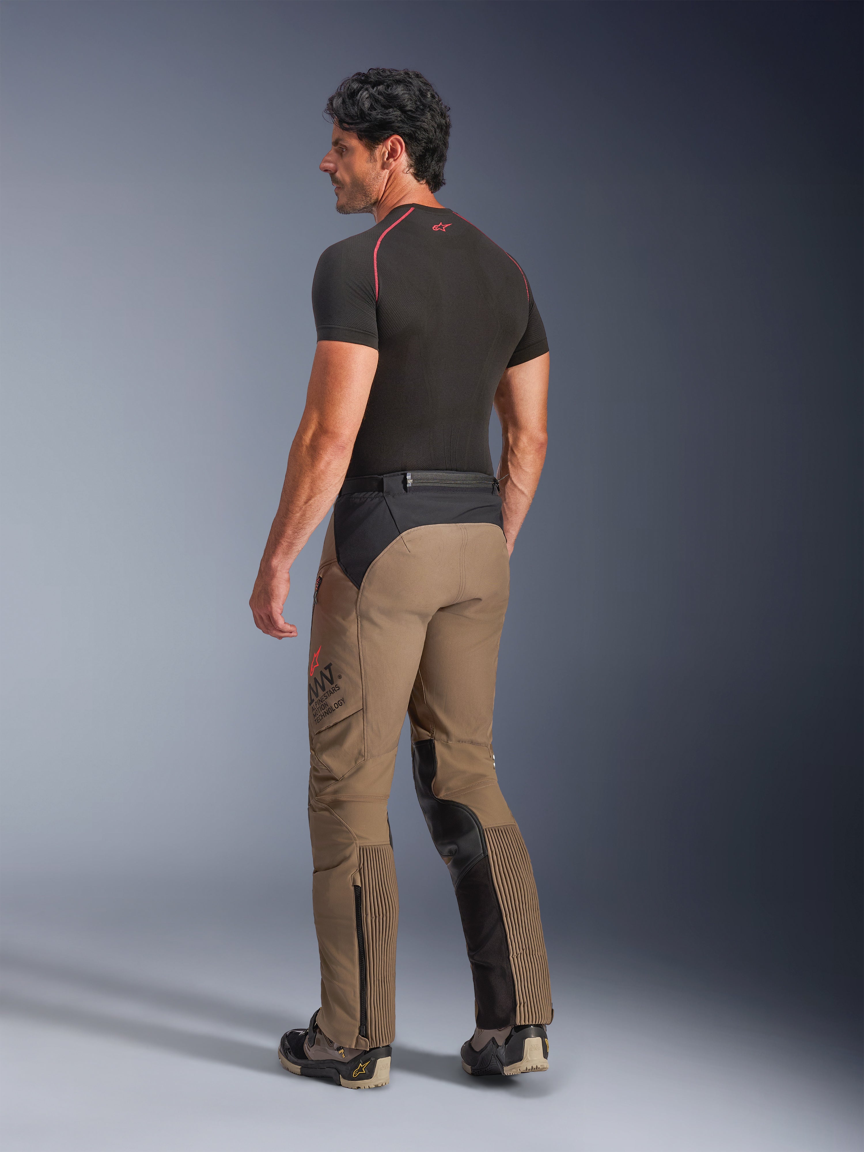 Pantaloni Alpinestars AMT-8 Stretch Drystar® XF in Walnut Black, pantaloni da moto adventure-touring visti da una prospettiva posteriore di tre quarti, in tessuto marrone noce con pannelli neri su seduta e interno gamba, inserti elasticizzati a fisarmonica sui polpacci e marchio AMT laterale, indossati con base layer nero.