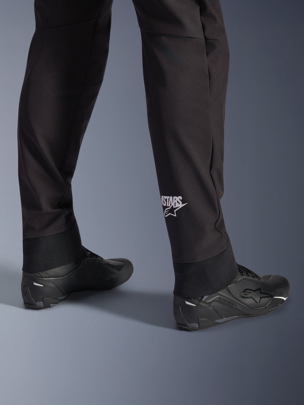 Alpinestars Aeroshell Pants, pantaloni da moto in softshell neri, dettaglio della gamba e dell'orlo elasticizzato, logo Alpinestars bianco sul polpaccio, abbinati a scarpe da moto nere, design crossover sportivo moderno