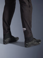 Alpinestars Aeroshell Pants, pantaloni da moto in softshell neri, dettaglio della gamba e dell'orlo elasticizzato, logo Alpinestars bianco sul polpaccio, abbinati a scarpe da moto nere, design crossover sportivo moderno