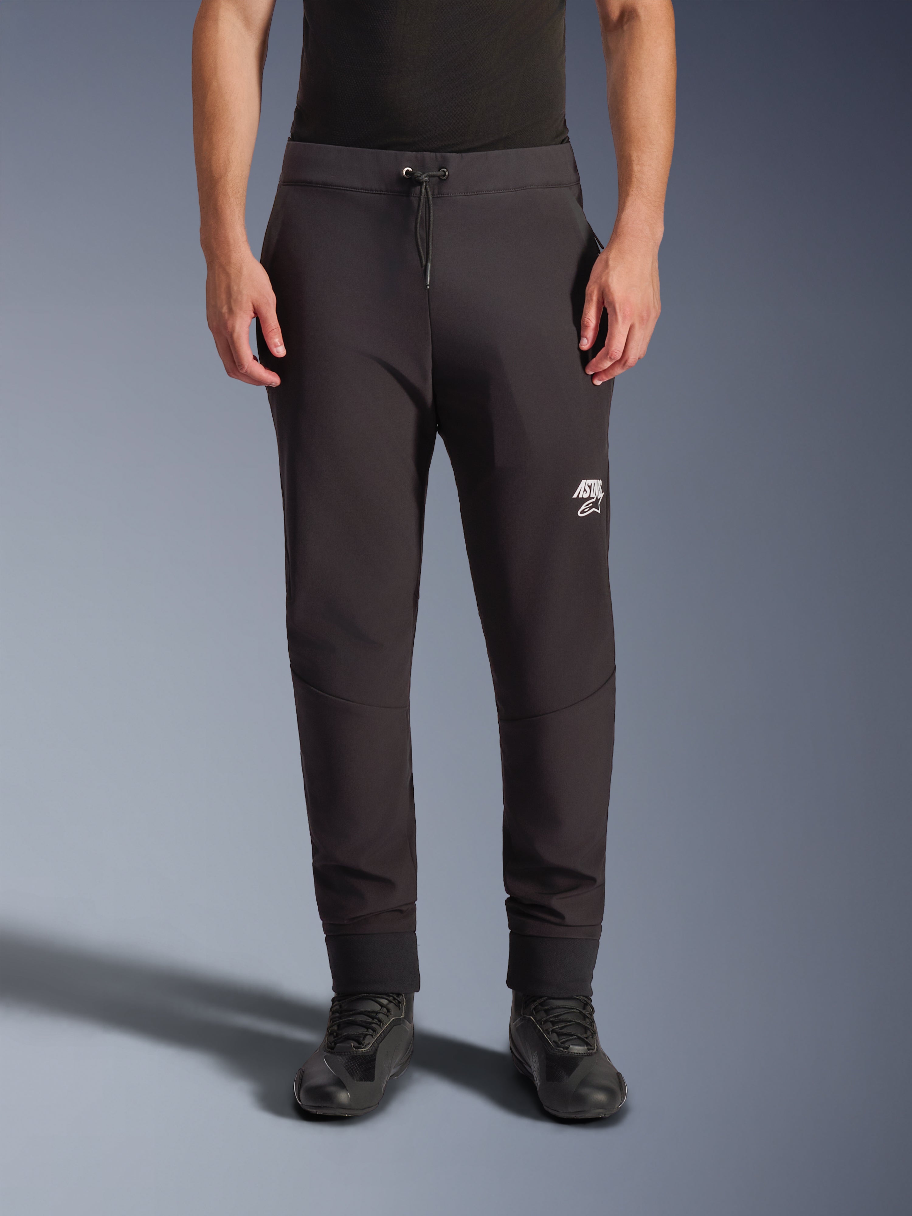 Alpinestars Aeroshell Pants, pantaloni da moto in softshell nero, vestibilità atletica stile jogger con coulisse e polsini elastici, logo Astars bianco, indossati con scarpe da guida nere, abbigliamento crossover moderno per uso stradale e casual