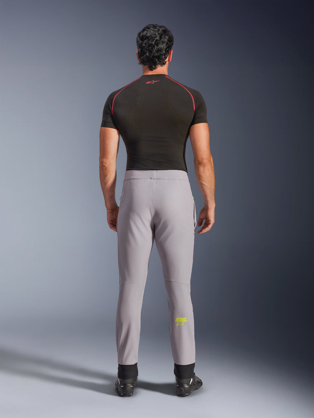 Alpinestars Aeroshell Pants, grigio medio con logo giallo fluo, vista posteriore di pantaloni da moto softshell sportivi crossover, indossati con maglia a compressione nera e scarpe da guida, con vita elastica e silhouette affusolata