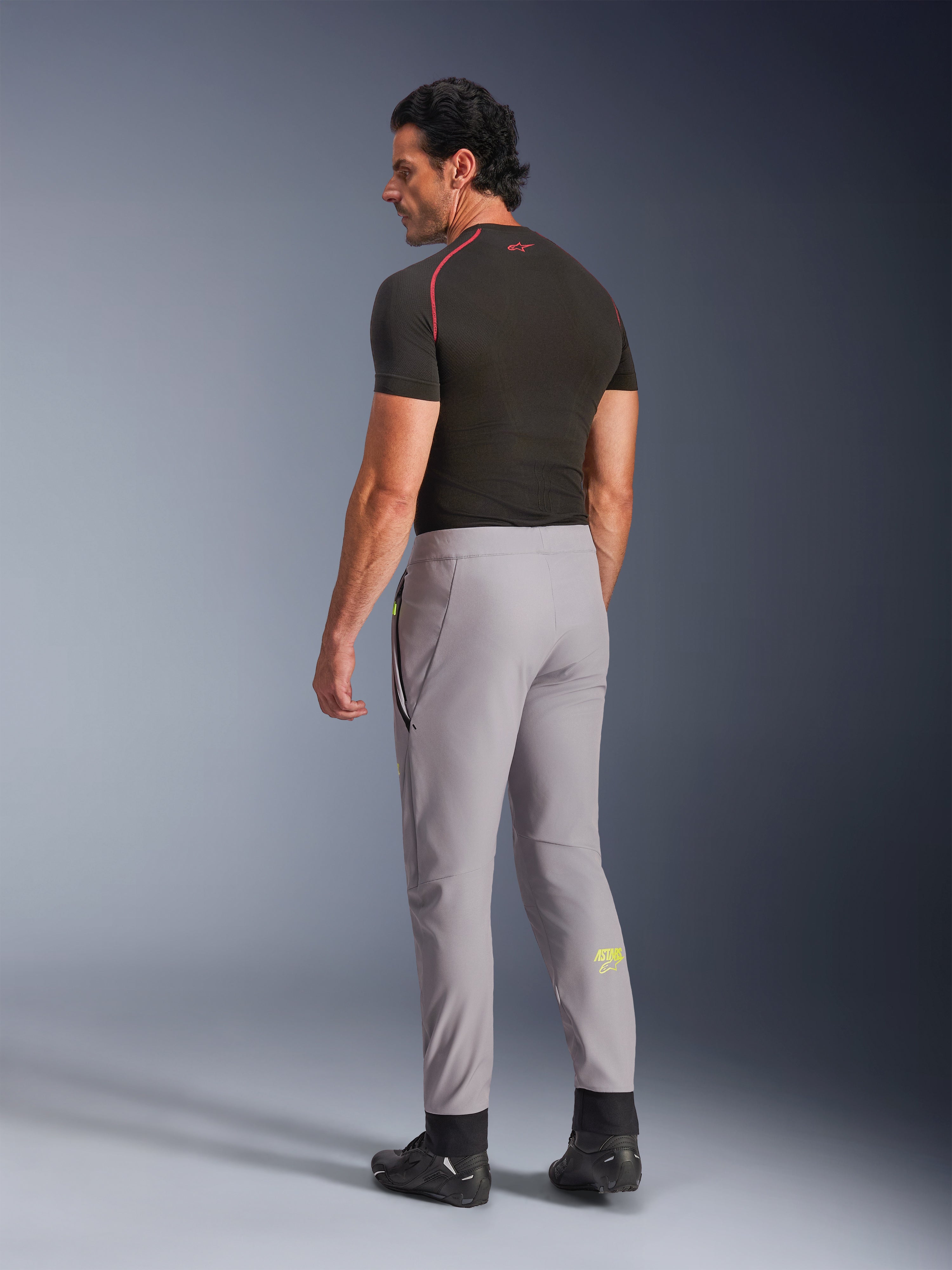 Alpinestars Pantaloni Aeroshell, Mid Gray Yellow Fluo, vista posteriore di un modello che indossa pantaloni da moto in softshell elasticizzato, logo Astars giallo sul polpaccio sinistro, abbinati a un base layer nero e scarpe tecniche per una silhouette sportiva moderna.